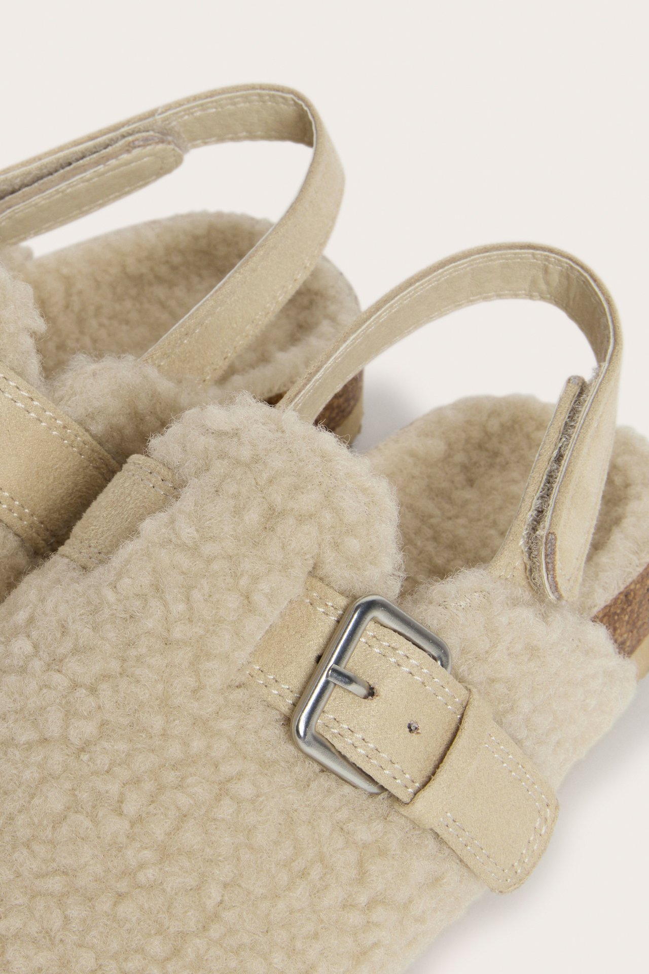 Lähikuva beigestä pörrötohvelista, jossa mokkanahkainen hihna, metallisolki ja takahihna.