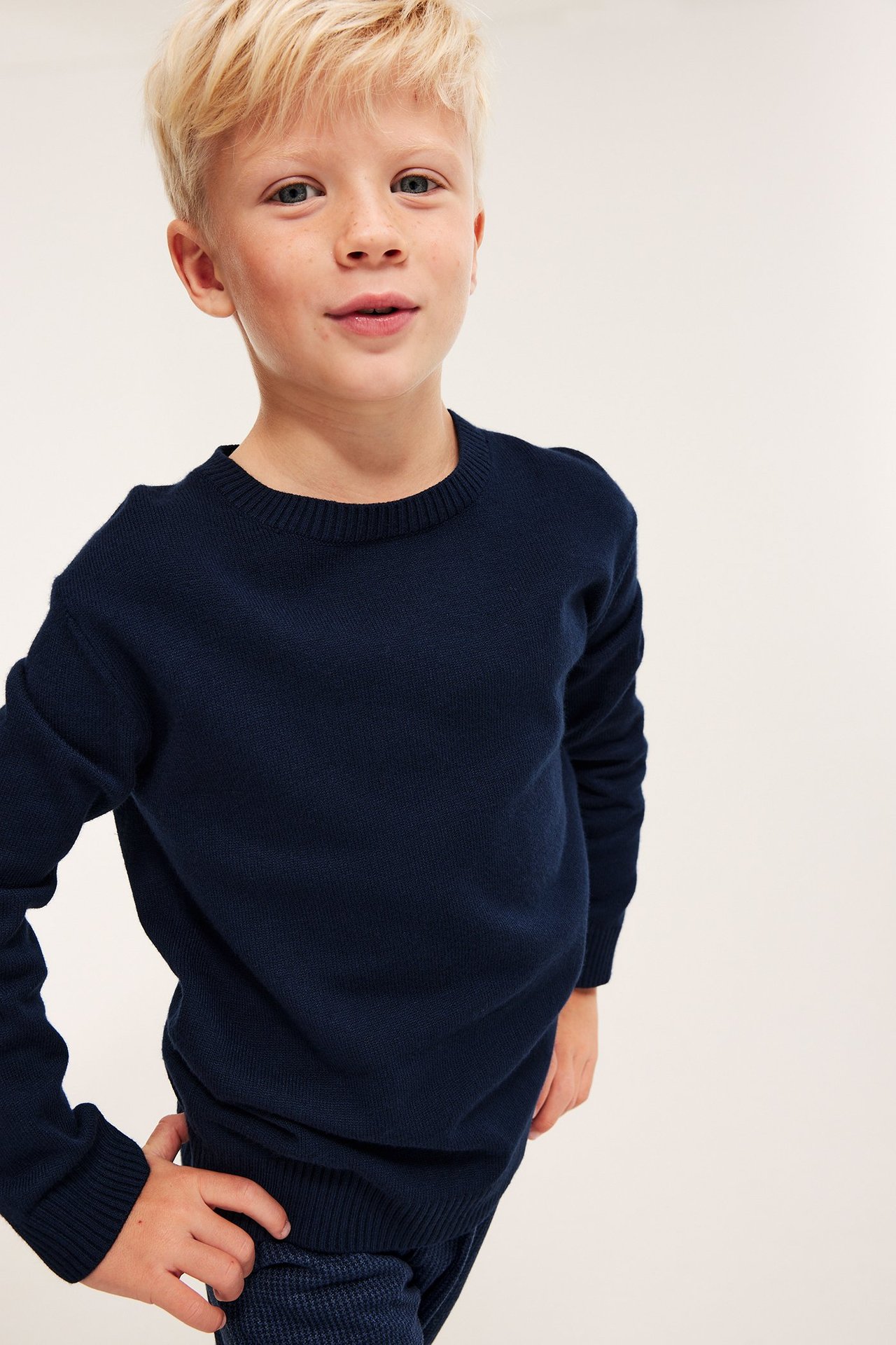 Framifrån, ett barn i mörkblå enfärgad stickad tröja med ribbade detaljer och mörka mönstrade byxor.