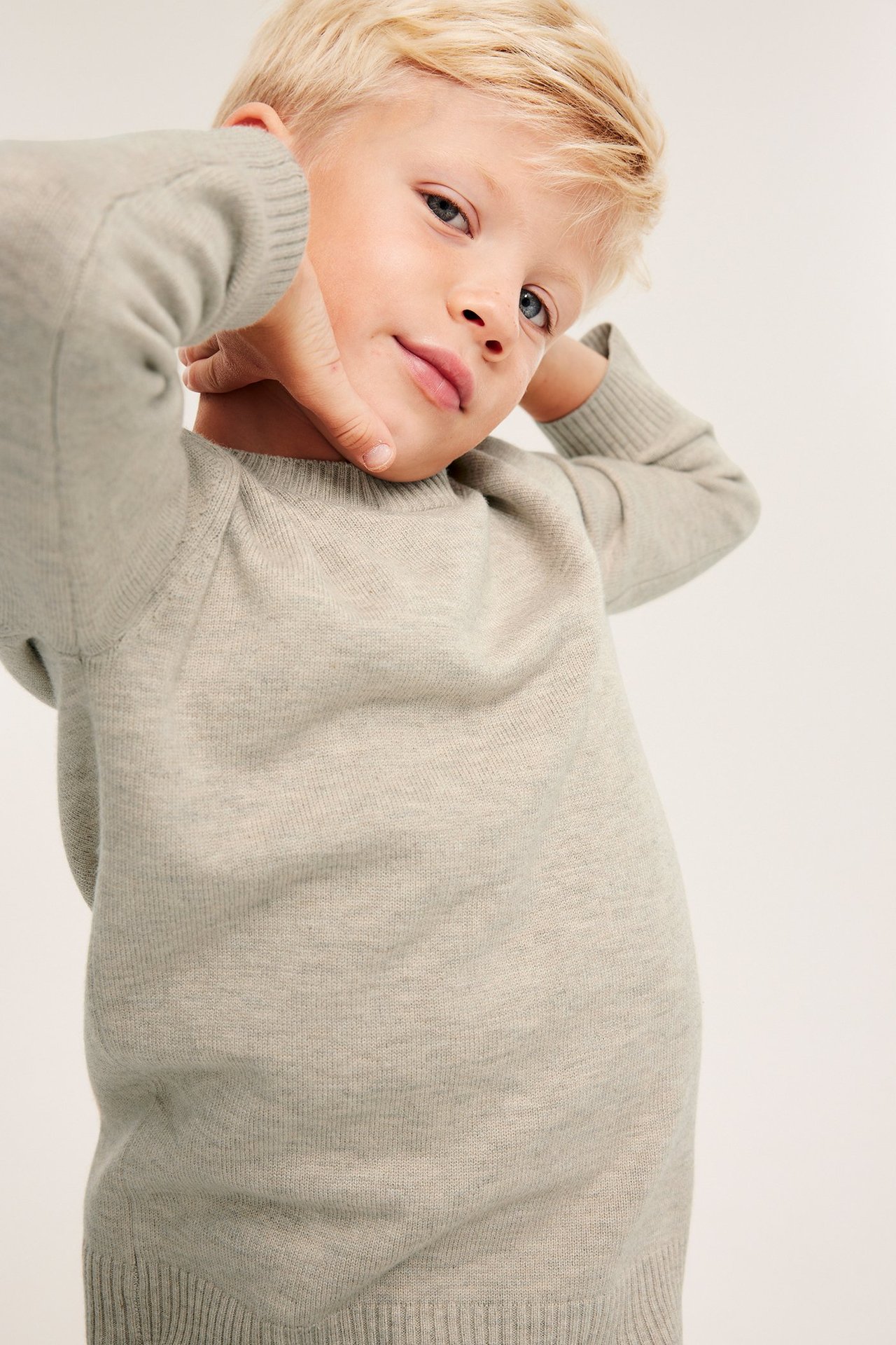 Frontvy: Barn i en ljusbeige melange stickad tröja med ribbade muddar och nederkant, händerna bakom huvudet.