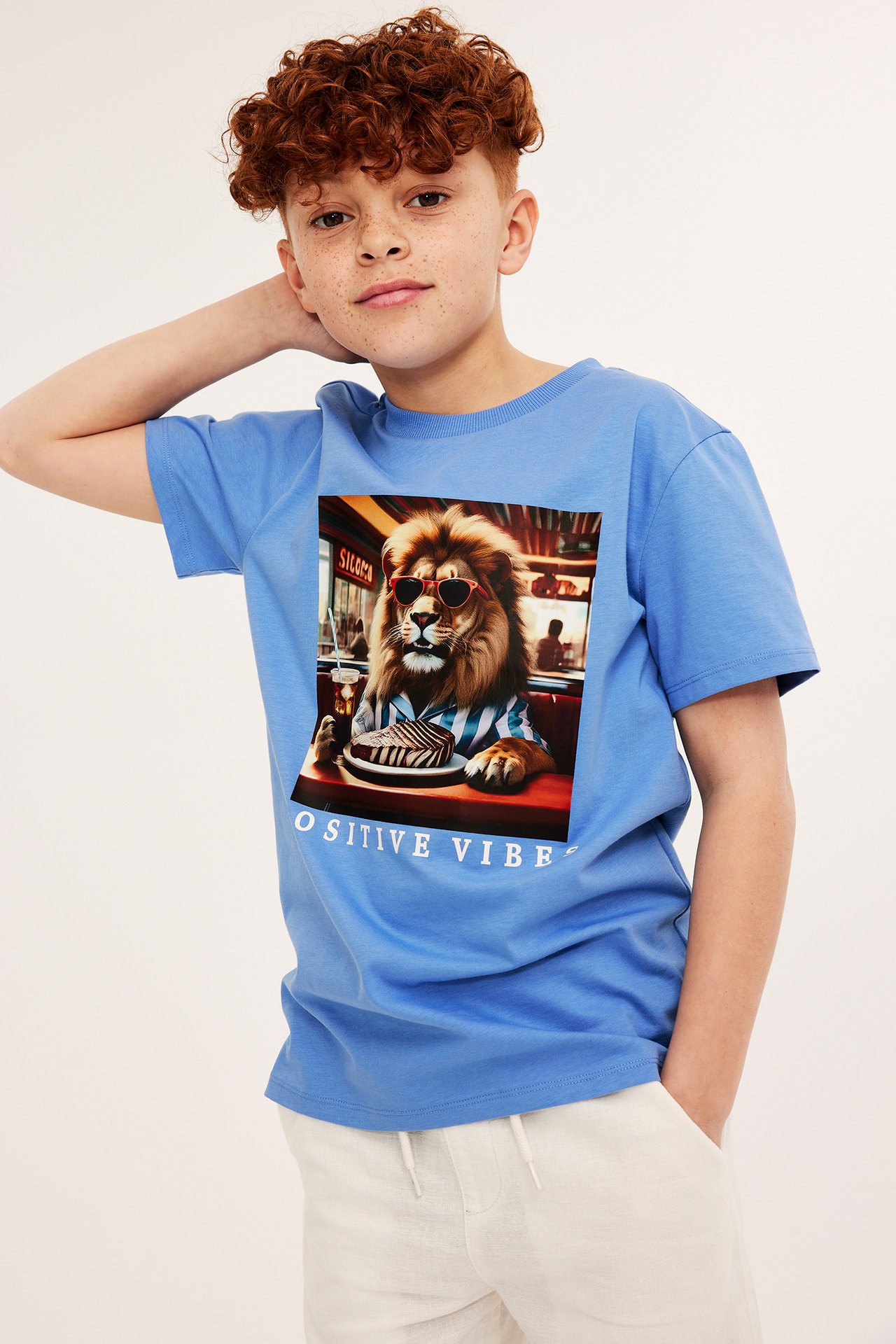 Framifrån, ett barn i en blå bomulls-T-shirt med lejontryck och ljusbeige shorts.