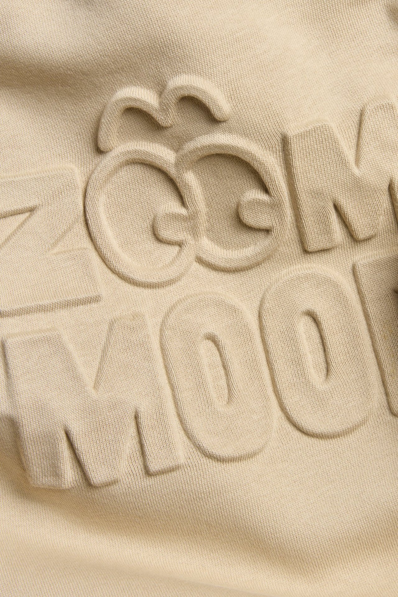 Nærbilde av en beige genser med et 3D-preget trykk foran.