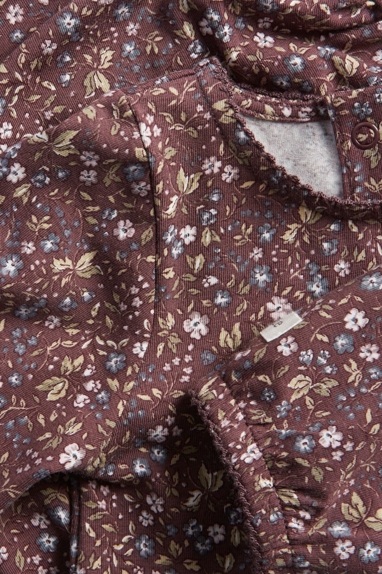 Zbliżenie na liliowe bawełniane body niemowlęce w kwiaty, z bufiastymi rękawami, wykończeniem picot i zatrzaskami.