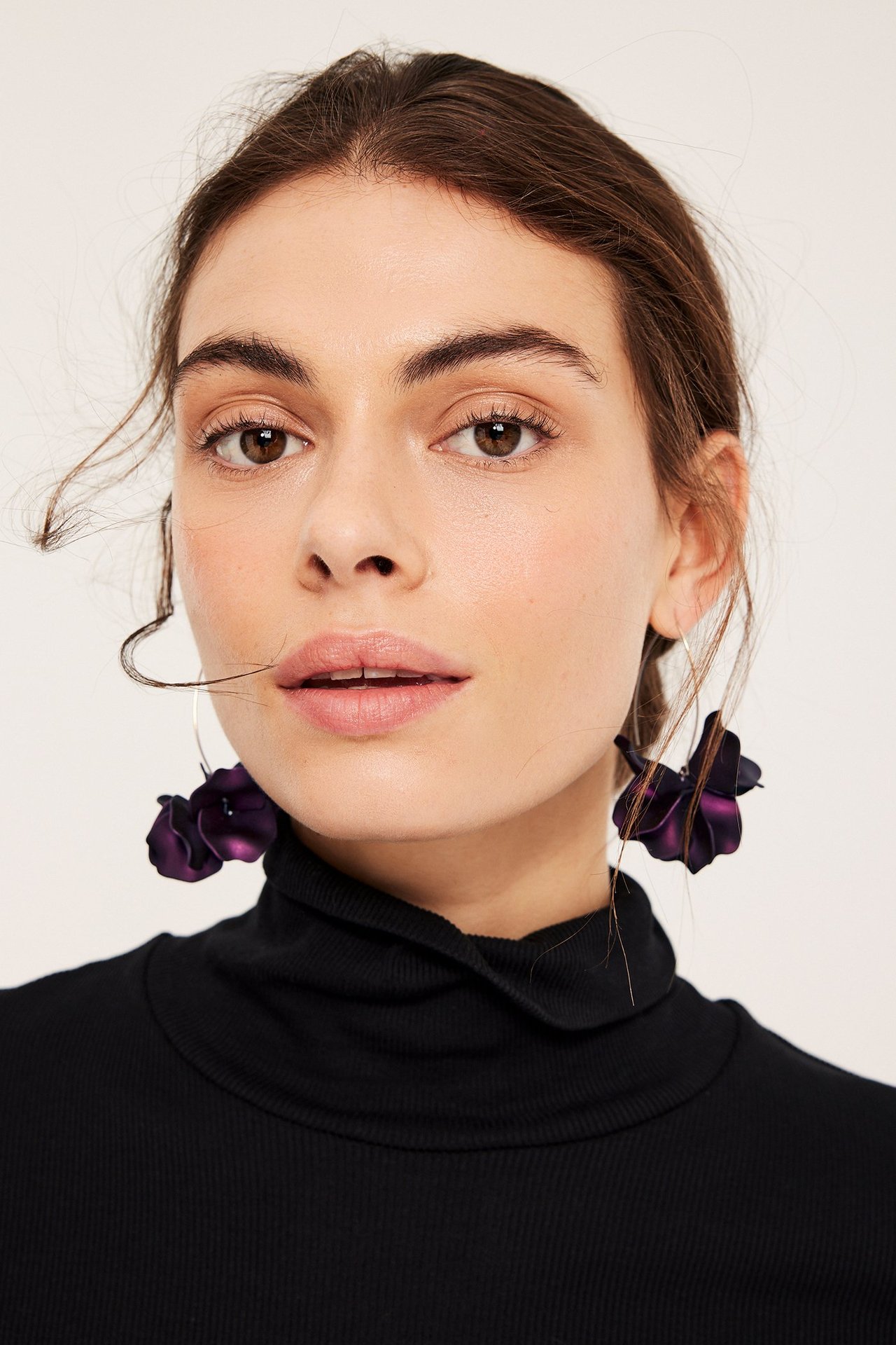 Zbliżenie na kobietę z przodu, noszącą długie śliwkowe kolczyki-kwiaty i czarny prążkowany golf.