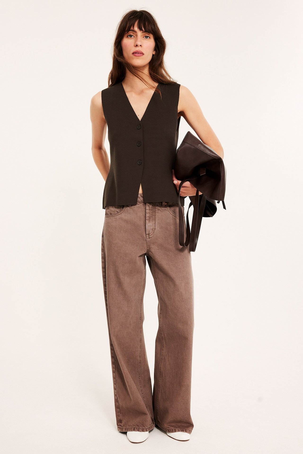 Framifrån: kvinna i mörkbrun stickad knäppt väst, bruna vida byxor, håller en mörkbrun handväska.