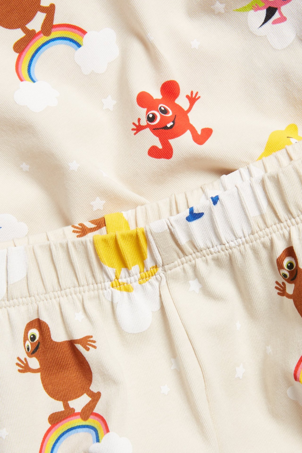 Zbliżenie na jasnobeżową piżamę dziecięcą z bawełny z kolorowym wzorem Babblarna, tęcz, chmur i gwiazd.