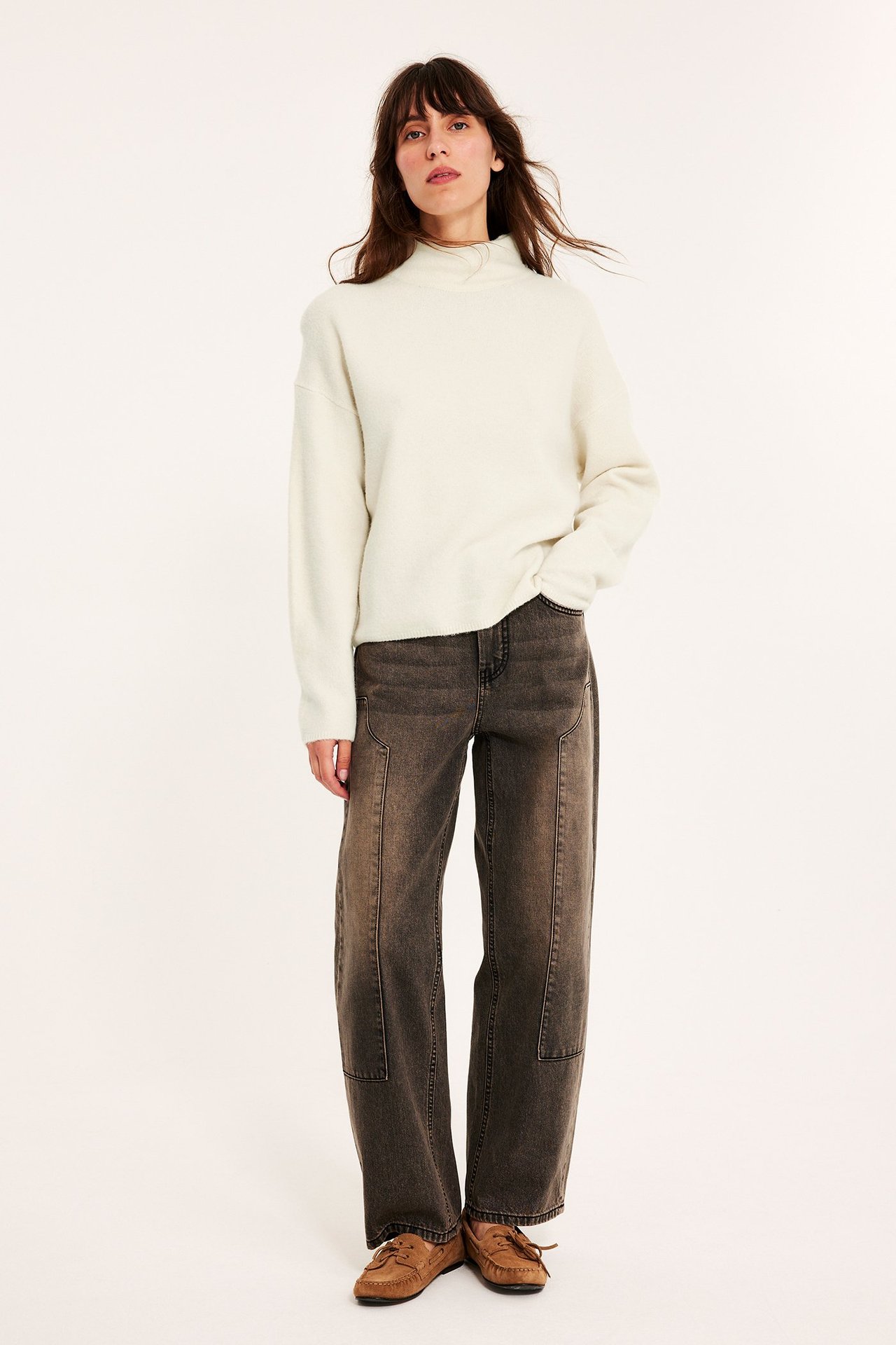 Frontbilde av en kvinne i lys beige strukturert høyhalset genser, mørkegrå vide jeans og brune mokkasiner.