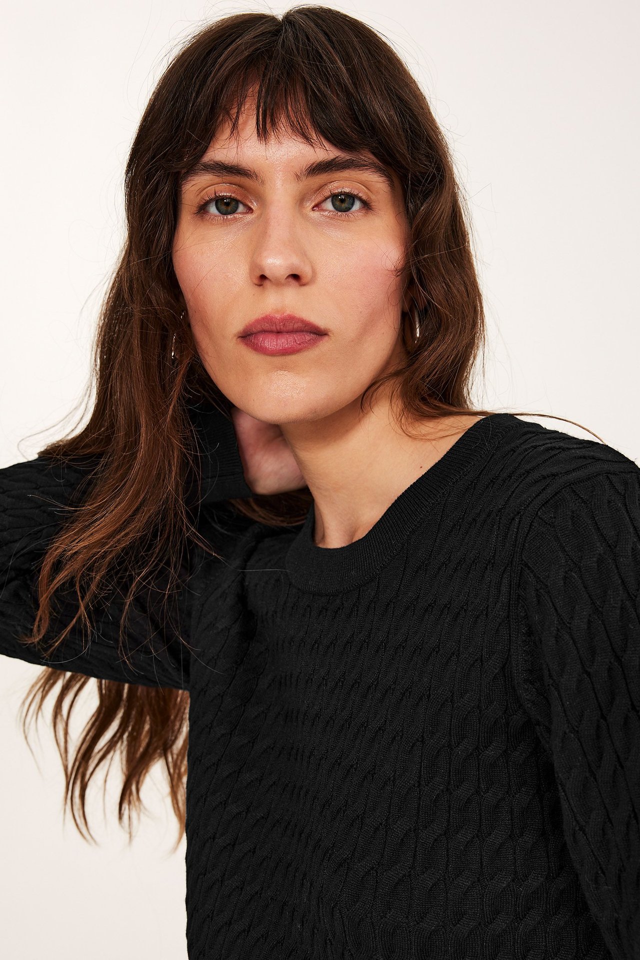 Nærbilde av en kvinne i svart kabelstrikket genser med rund hals og små sølvfargede ringøreringer.