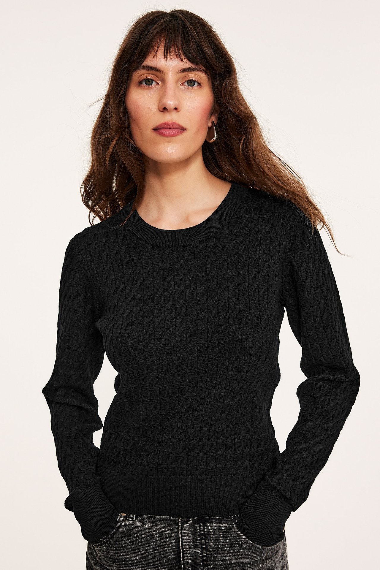 Forfra: kvinne i svart kabelstrikket genser, mørke jeans og små gullfargede øreringer.