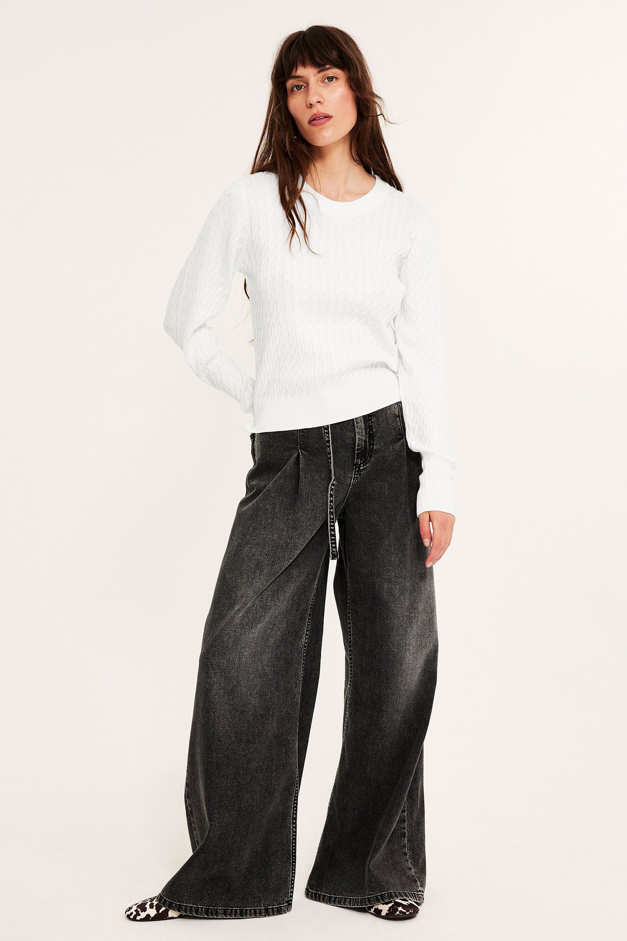 Frontbilde av en kvinne i en offwhite kabelstrikket genser, mørkegrå vide jeans og dyremønstrede flate sko.