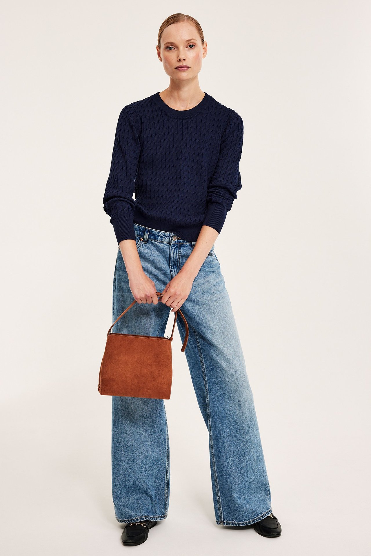 Etunäkymä naisesta tummansinisessä palmikkoneuleessa, vaaleansinisissä farkuissa, ja ruskea mokkanahkalaukku.