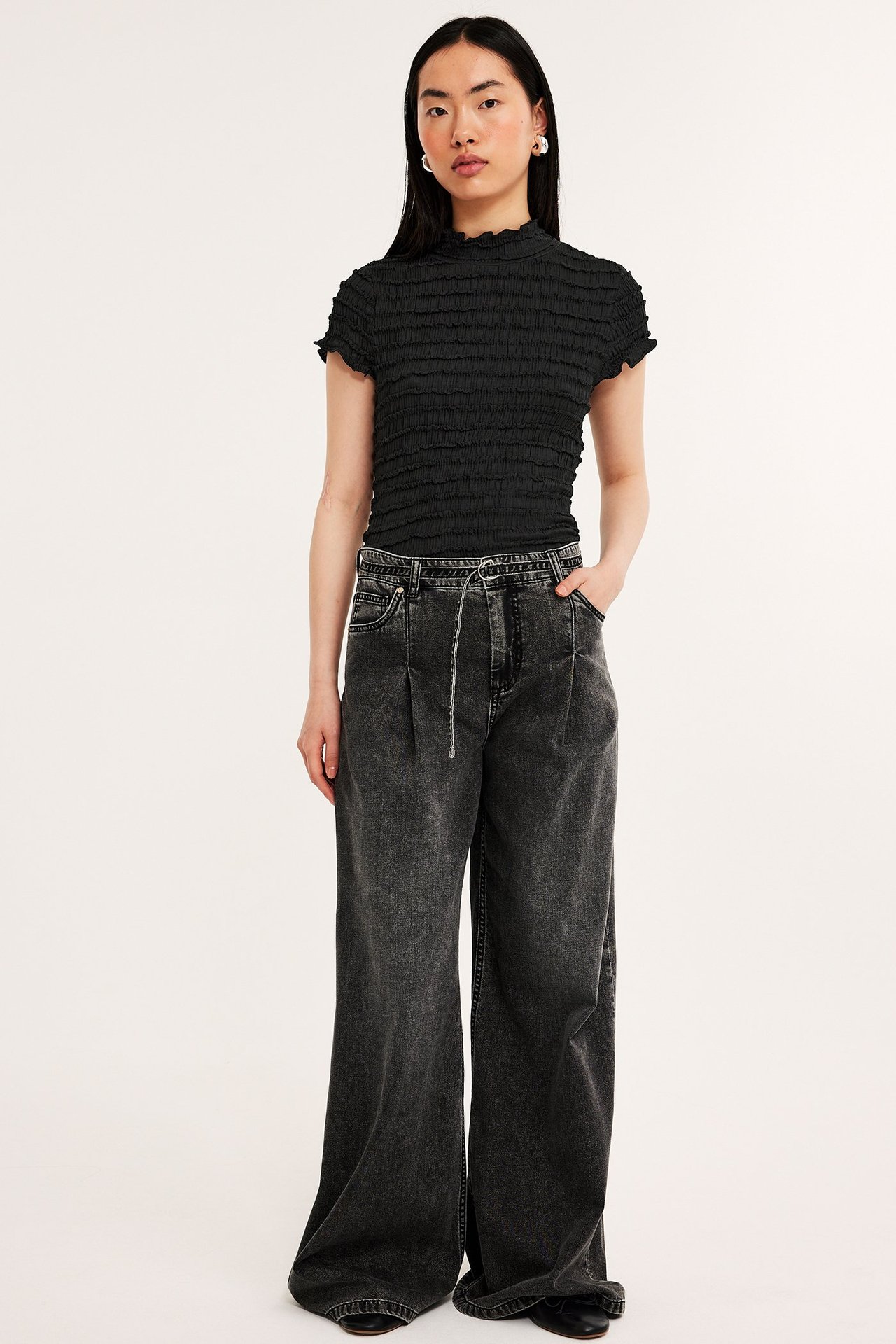 Framifrån: kvinna i svart strukturerad kortärmad topp, mörkgrå vida jeans och svarta skor.