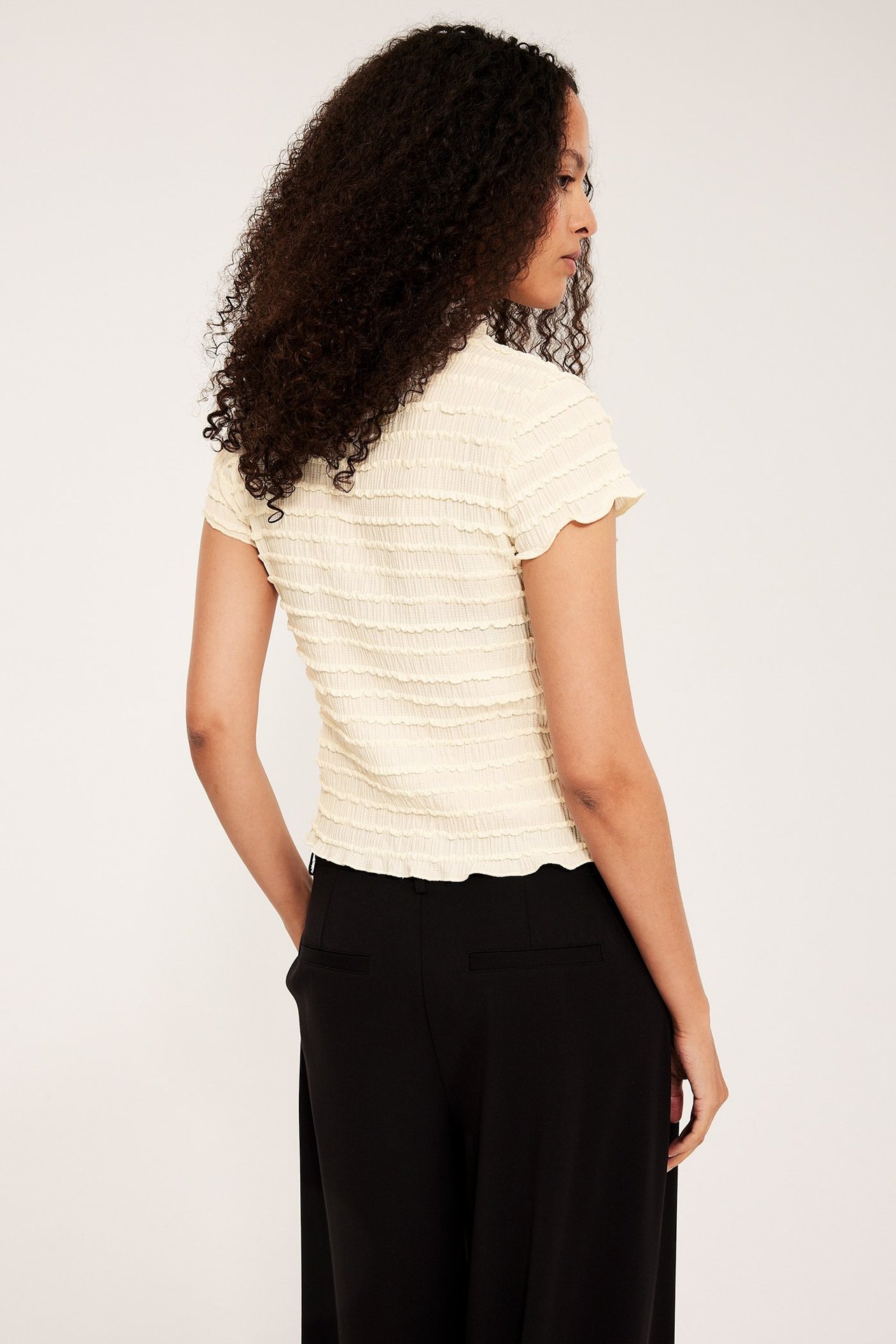 Widok z tyłu kobiety w kremowym, strukturalnym topie z krótkim rękawem i czarnych szerokich spodniach.