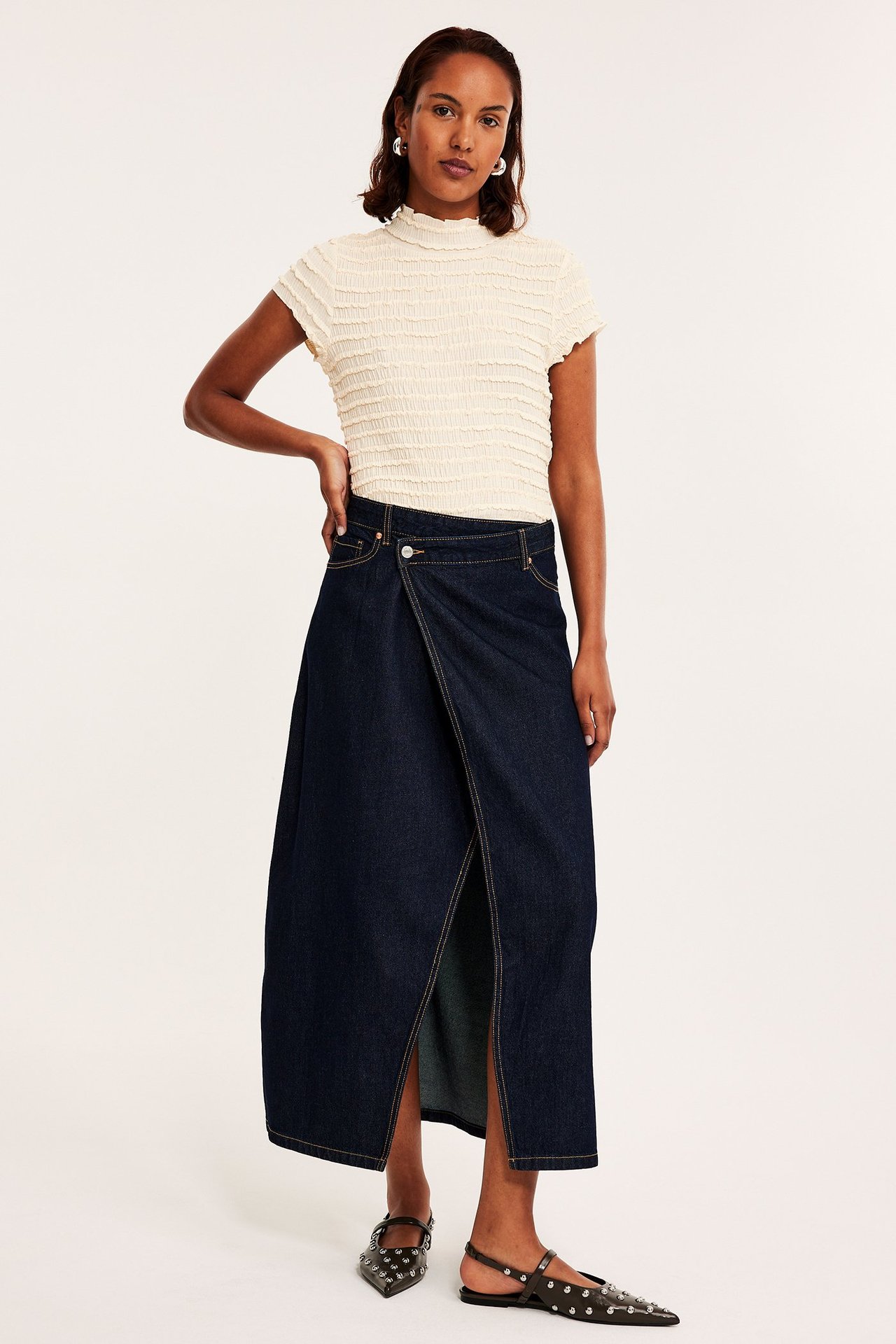 Framifrån: kvinna i offwhite strukturerad kortärmad topp, mörkblå omlott-jeanskjol och svarta nitprydda platta skor.