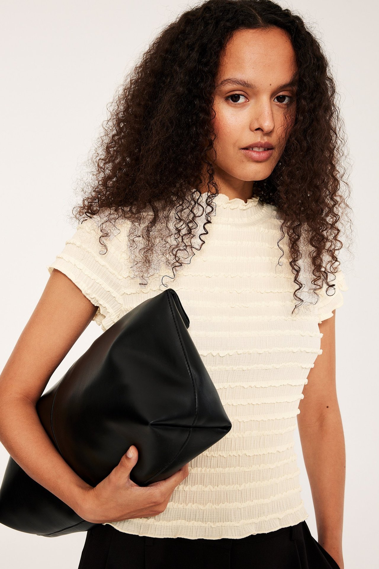 Kobieta od przodu w ecru, strukturalnym topie z krótkim rękawem i wysokim kołnierzem, trzyma czarną torebkę.