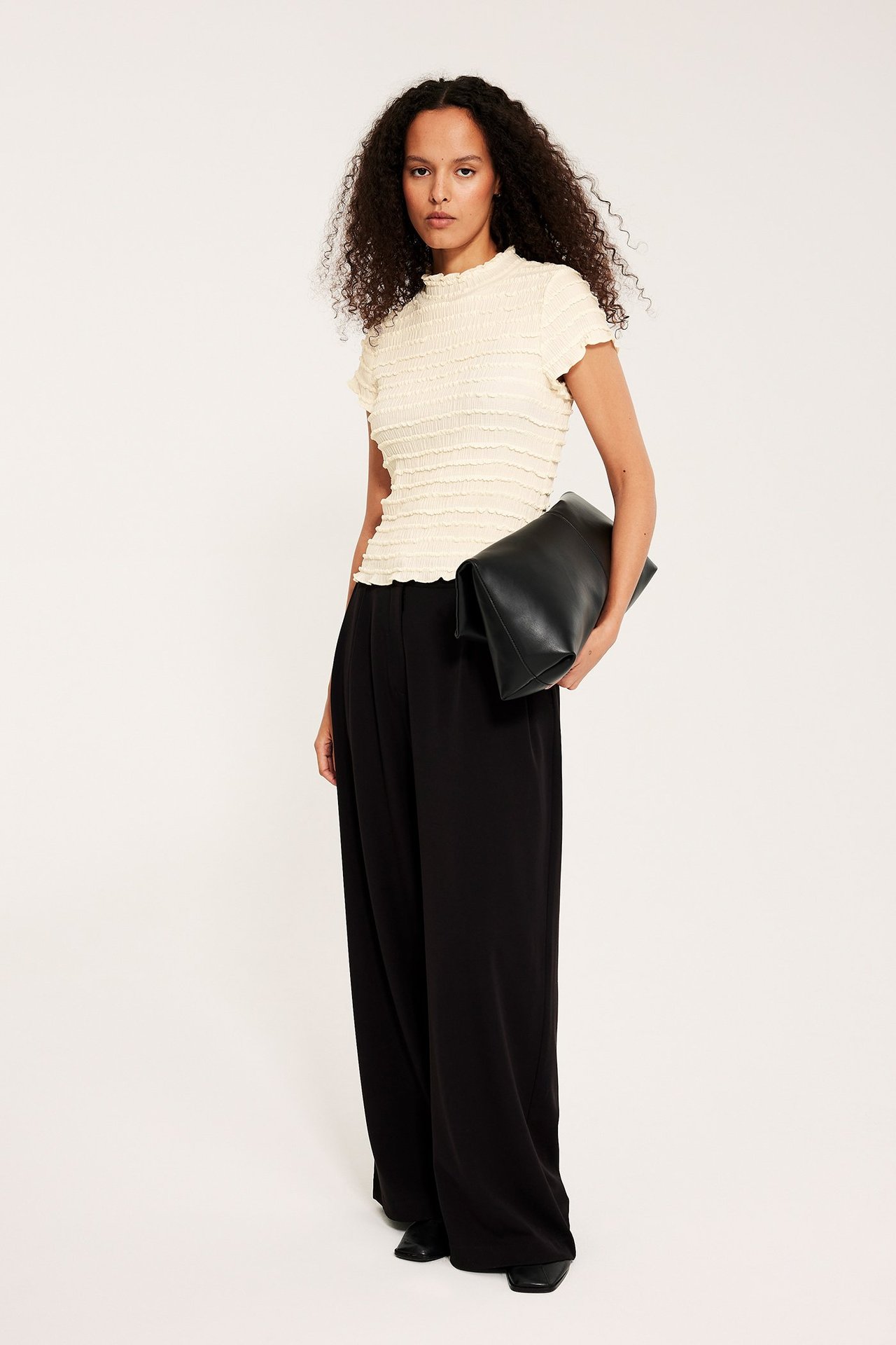 Frontbilde av kvinne i offwhite strukturert kortermet topp, svarte vide bukser, holder svart clutchveske.