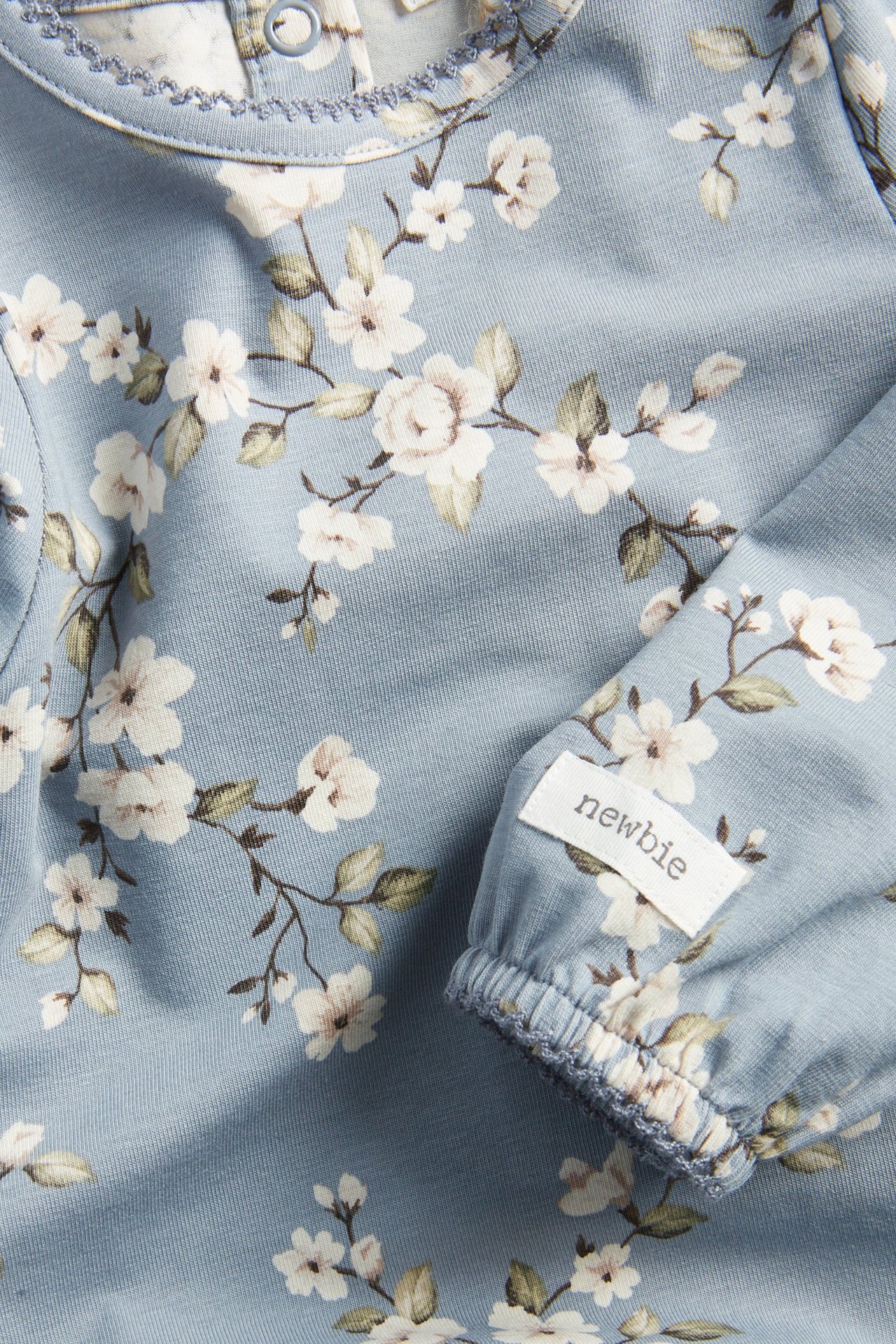 Zbliżenie na jasnoniebieskie, kwieciste body niemowlęce z długim rękawem, bufiastymi rękawami i wykończeniem picot.