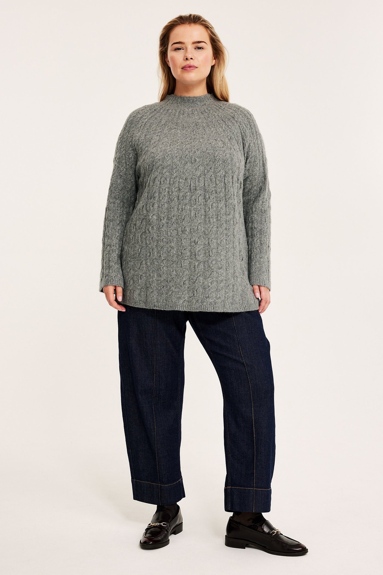 Edestä kuvattu nainen yllään harmaameleerattu palmikkoneule, tummansiniset leveät housut ja tummat loaferit.