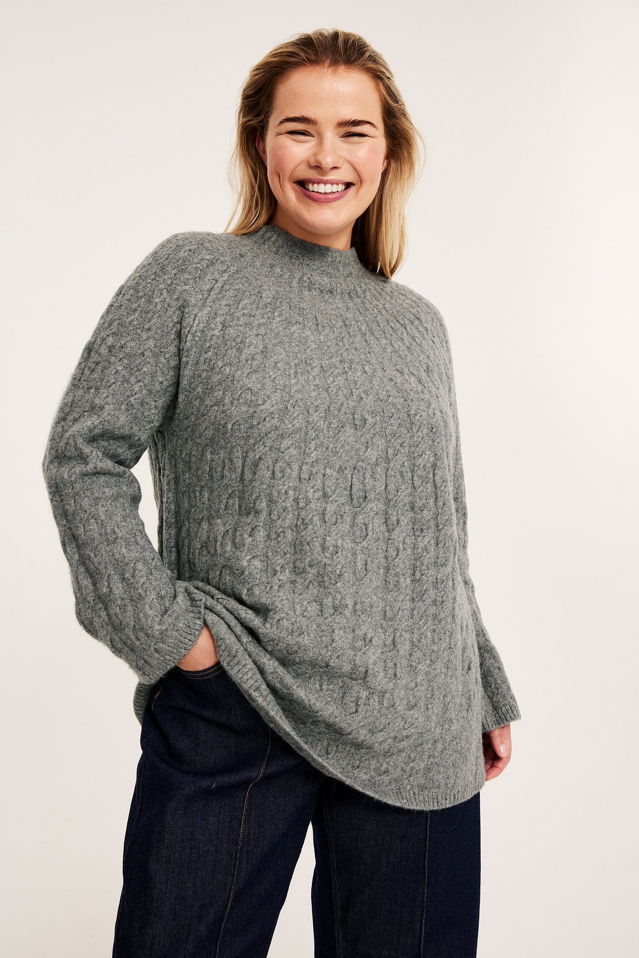 Edestä kuvattu hymyilevä nainen harmaassa palmikkoneuleisessa A-linjaisessa puserossa ja tummansinisissä farkuissa.