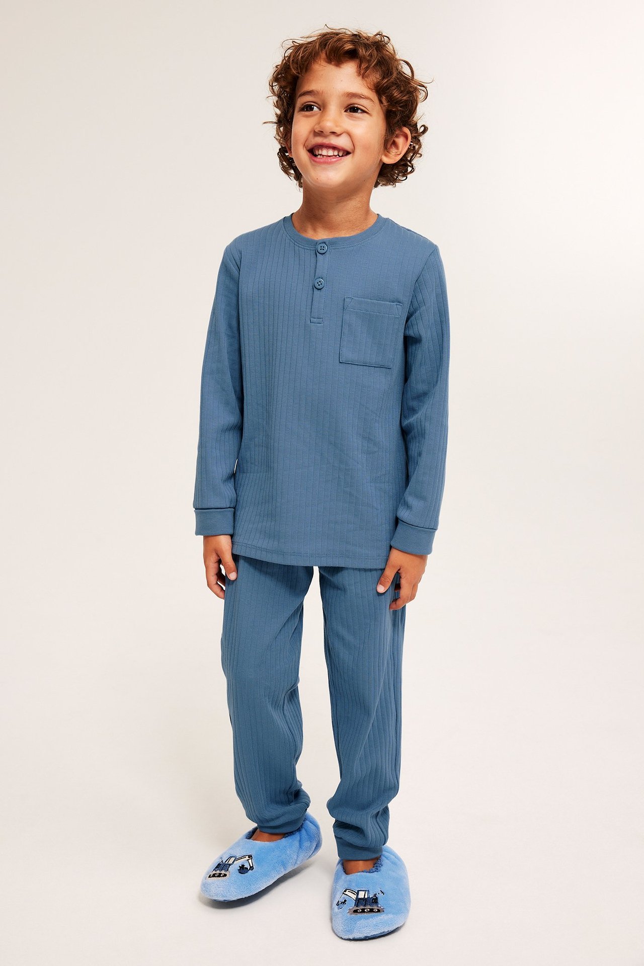 Edestä hymyilevä lapsi yllään sininen ribattu pyjama napeilla ja rintataskulla, sekä siniset tossut.