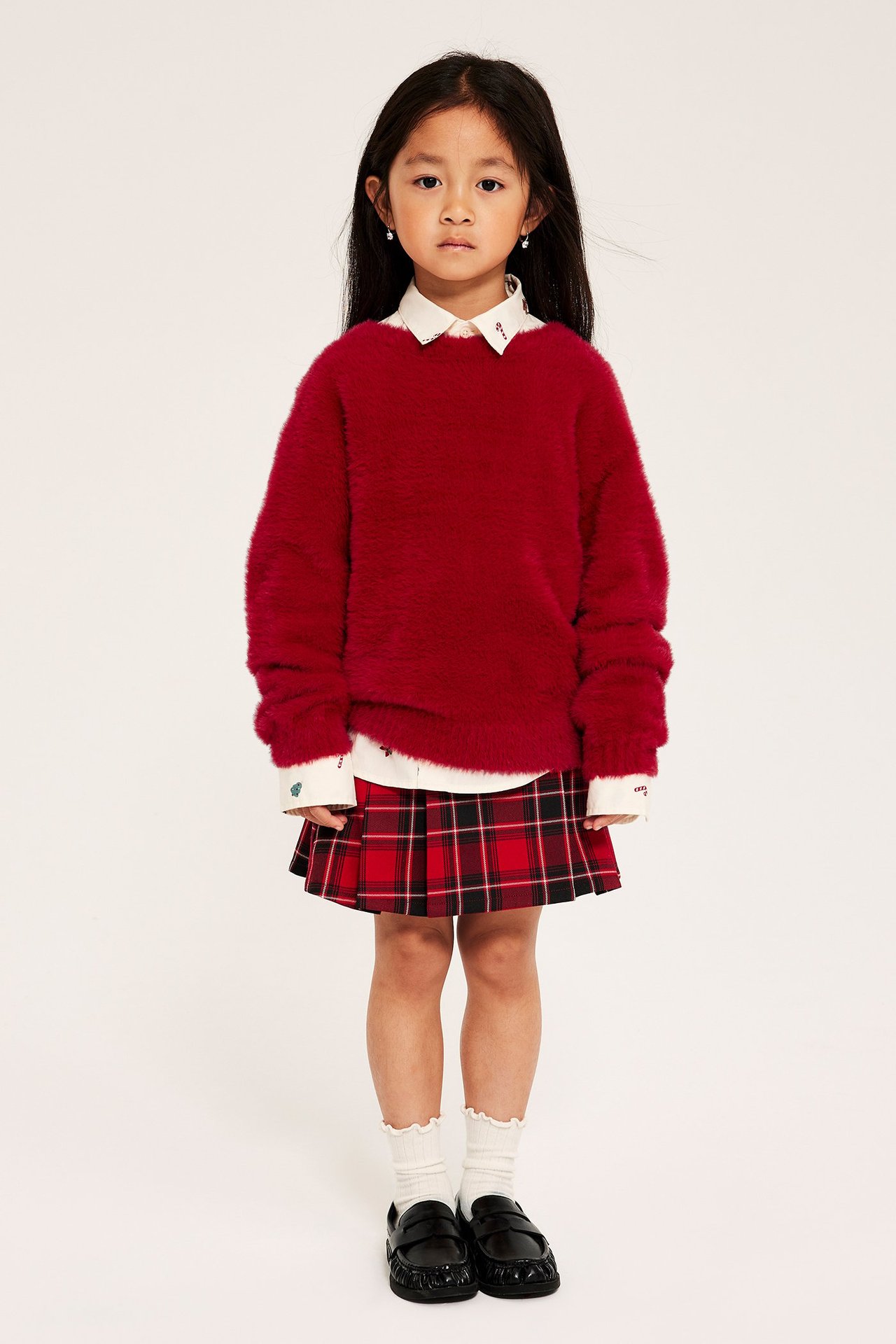 Dziewczynka w czerwonym, miękkim swetrze z feather yarn, z białą koszulą i czerwonym spódnicą w kratę.