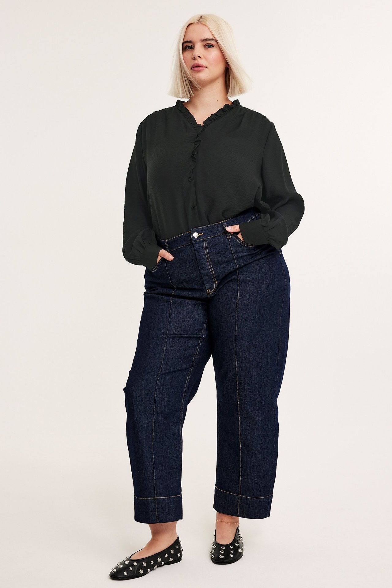 Framifrån: kvinna i svart volangblus, mörkblå vida jeans och svarta ballerinaskor med nitar.