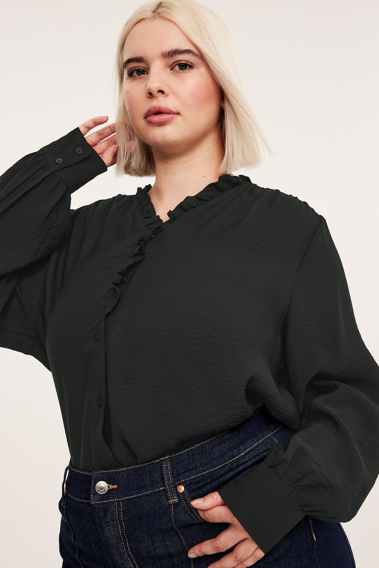 Närbild framifrån: kvinna i svart texturerad blus med volanghals och mörkblå jeans.