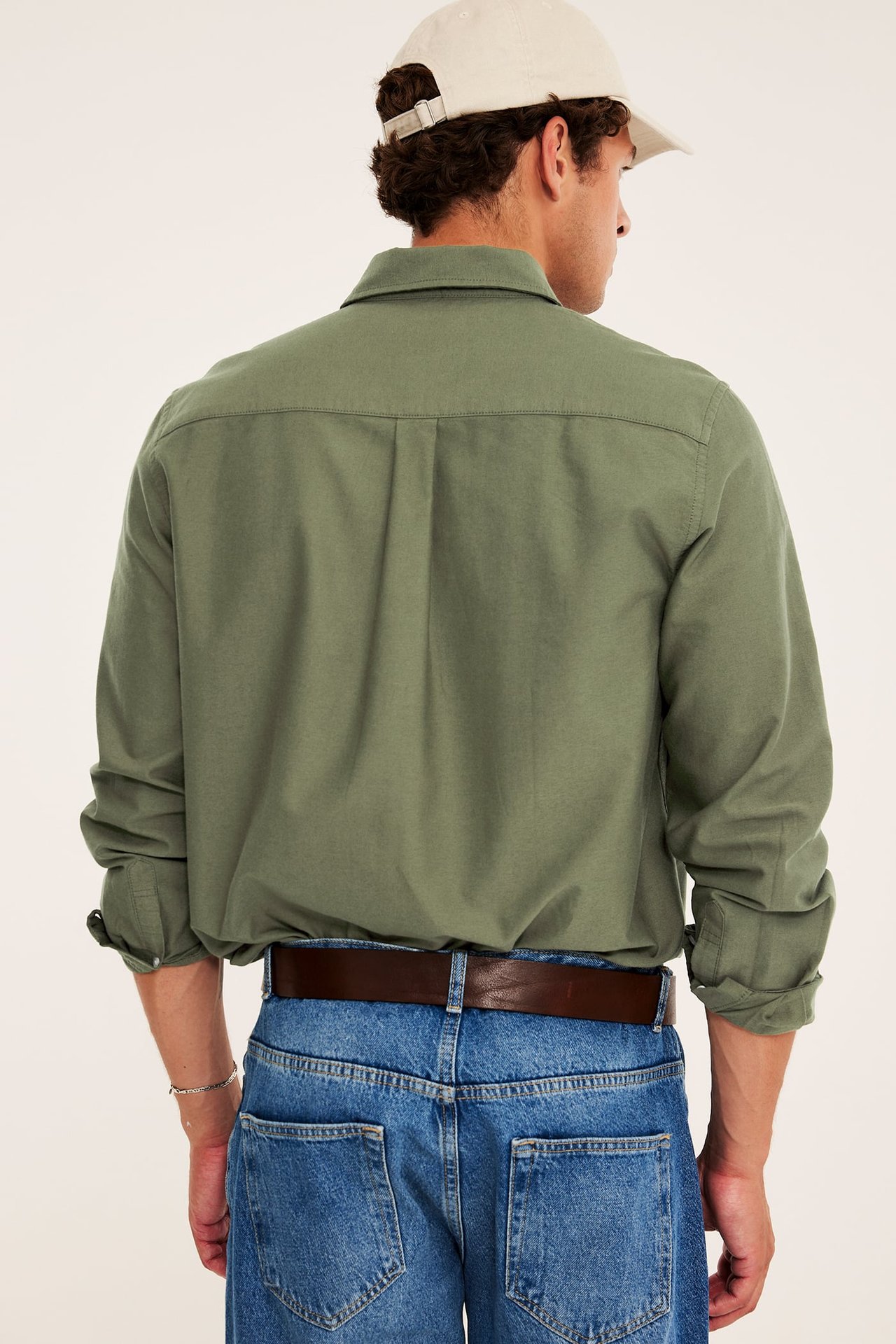Bakfra av mannlig modell iført grønn langermet skjorte, blå jeans, brunt belte og beige caps.