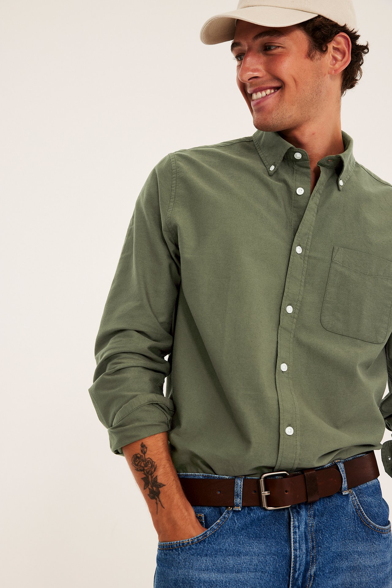 Framifrån: Vuxen man bär en olivgrön skjorta, blå jeans, brunt bälte och beige keps.