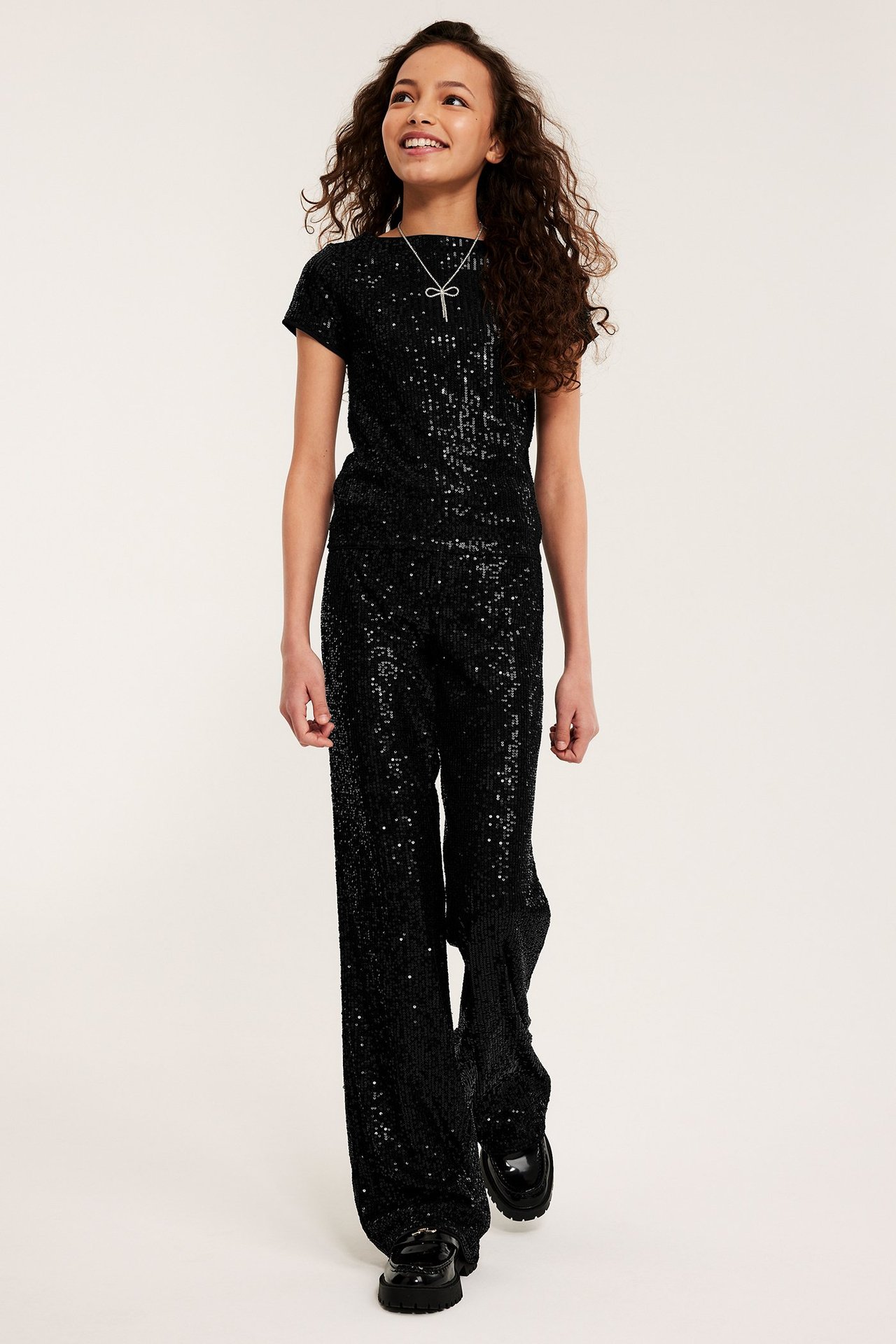 Framifrån: Barn i svart kortärmad paljettopp, matchande byxor, svarta loafers och ett silverhalsband.