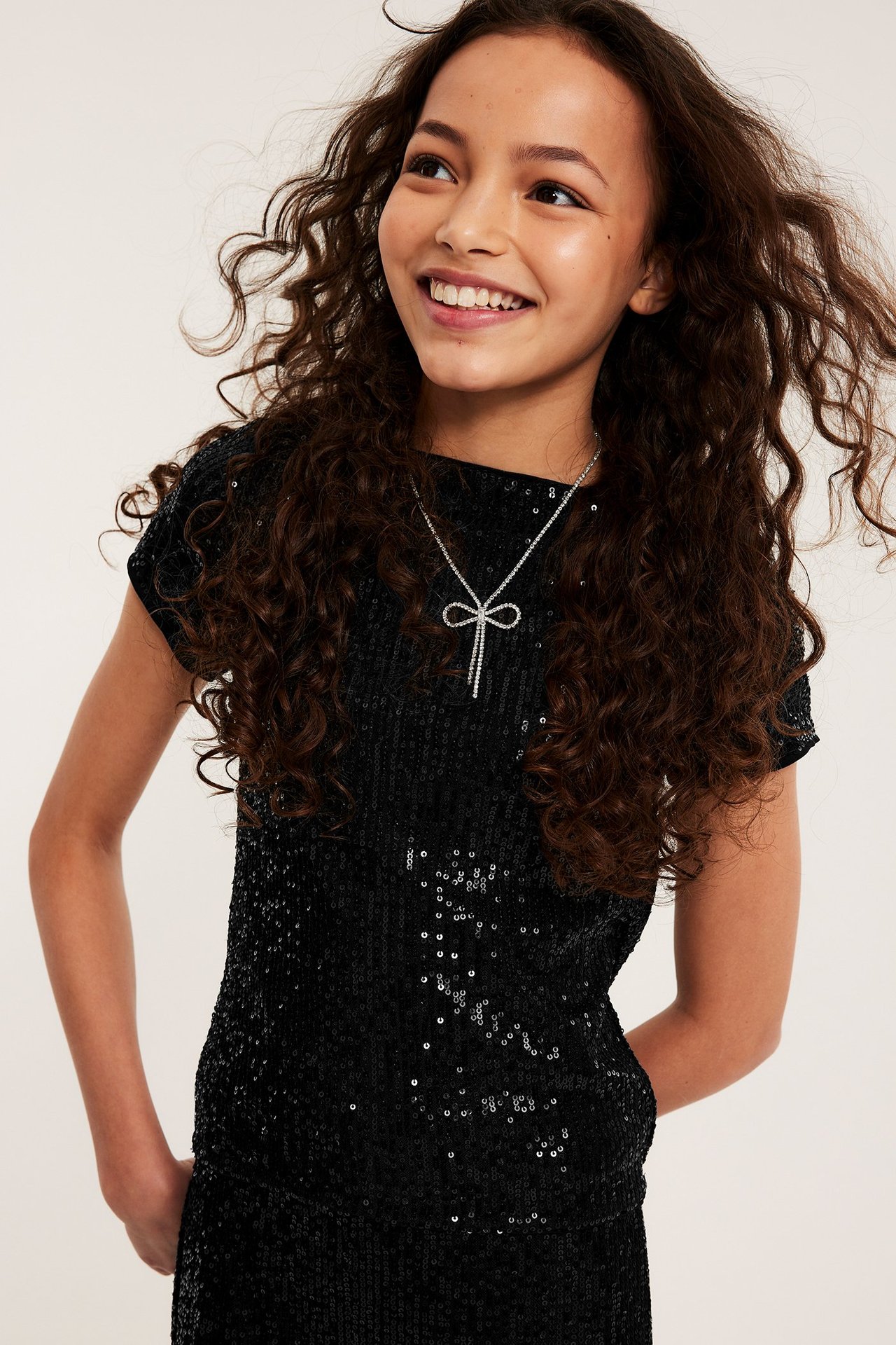 Frontvy av ett leende barn i en svart kortärmad paljettopp och ett silverfärgat rosetthalsband.