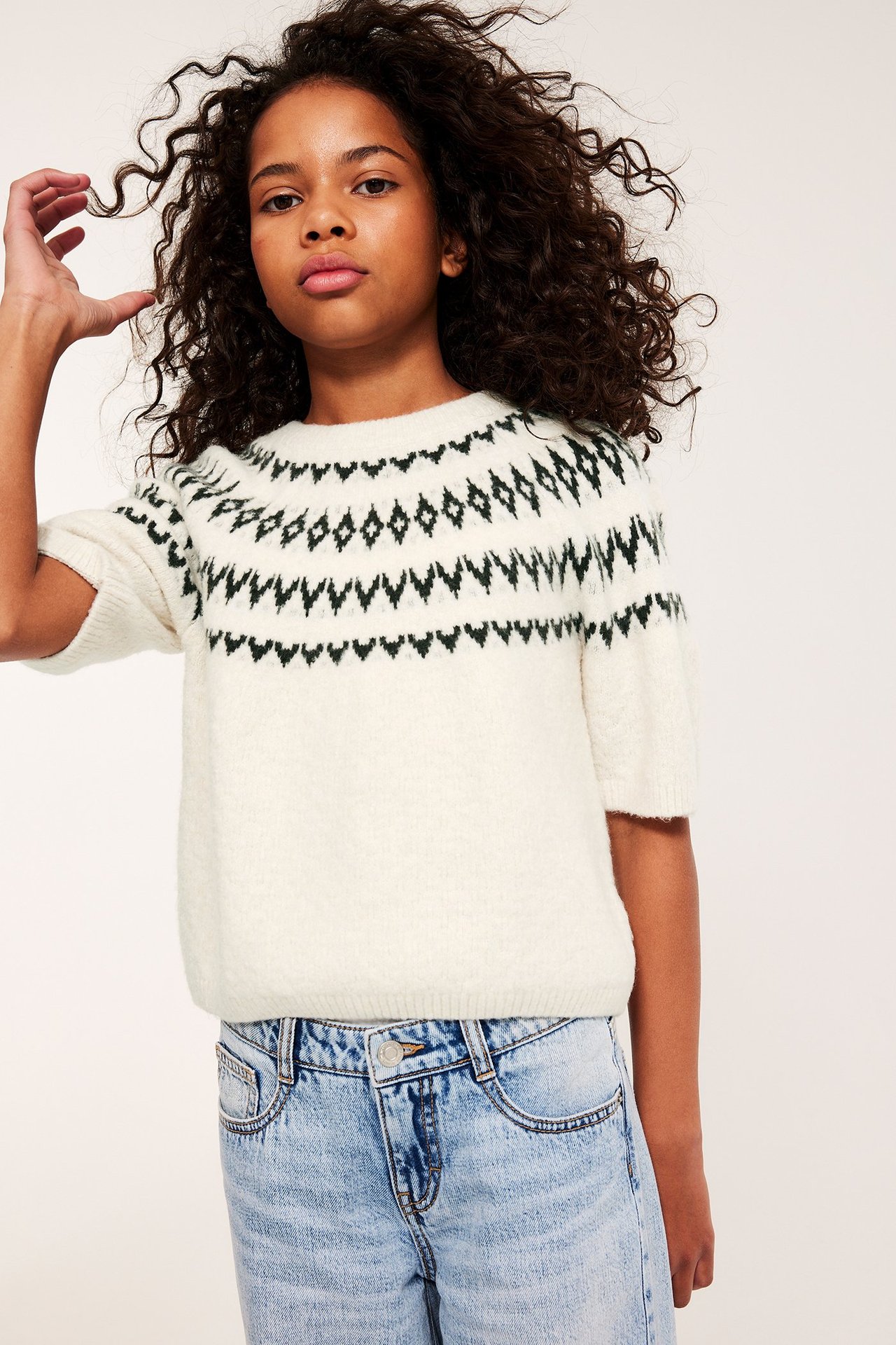 Framifrån, ett barn i en vit kortärmad stickad jacquardtröja och ljusblå tvättade jeans.