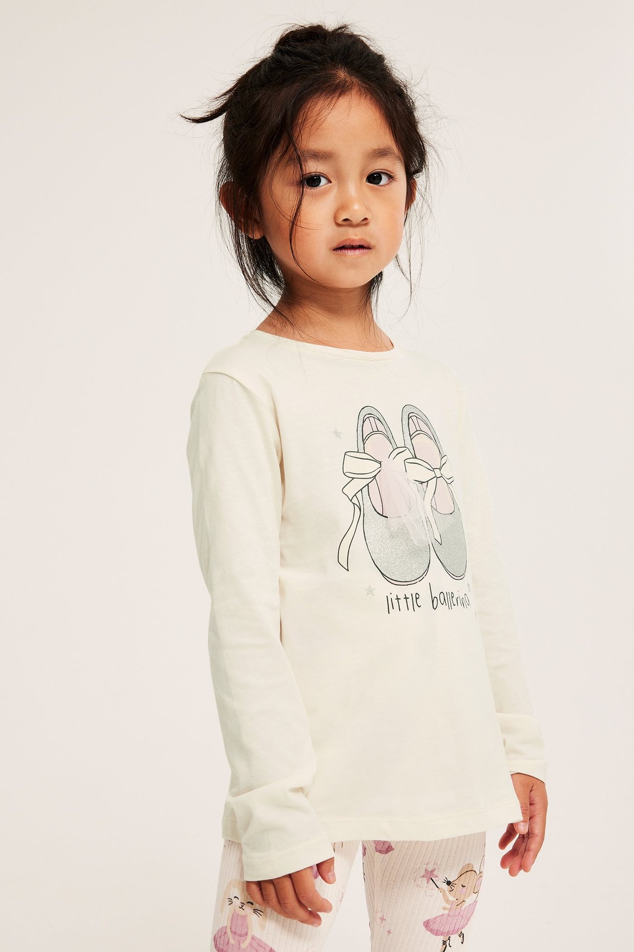 Edestäpäin lapsi yllään luonnonvalkoinen puuvillainen pitkähihainen toppi balettikenkäprintillä ja kuvioidut leggingsit.