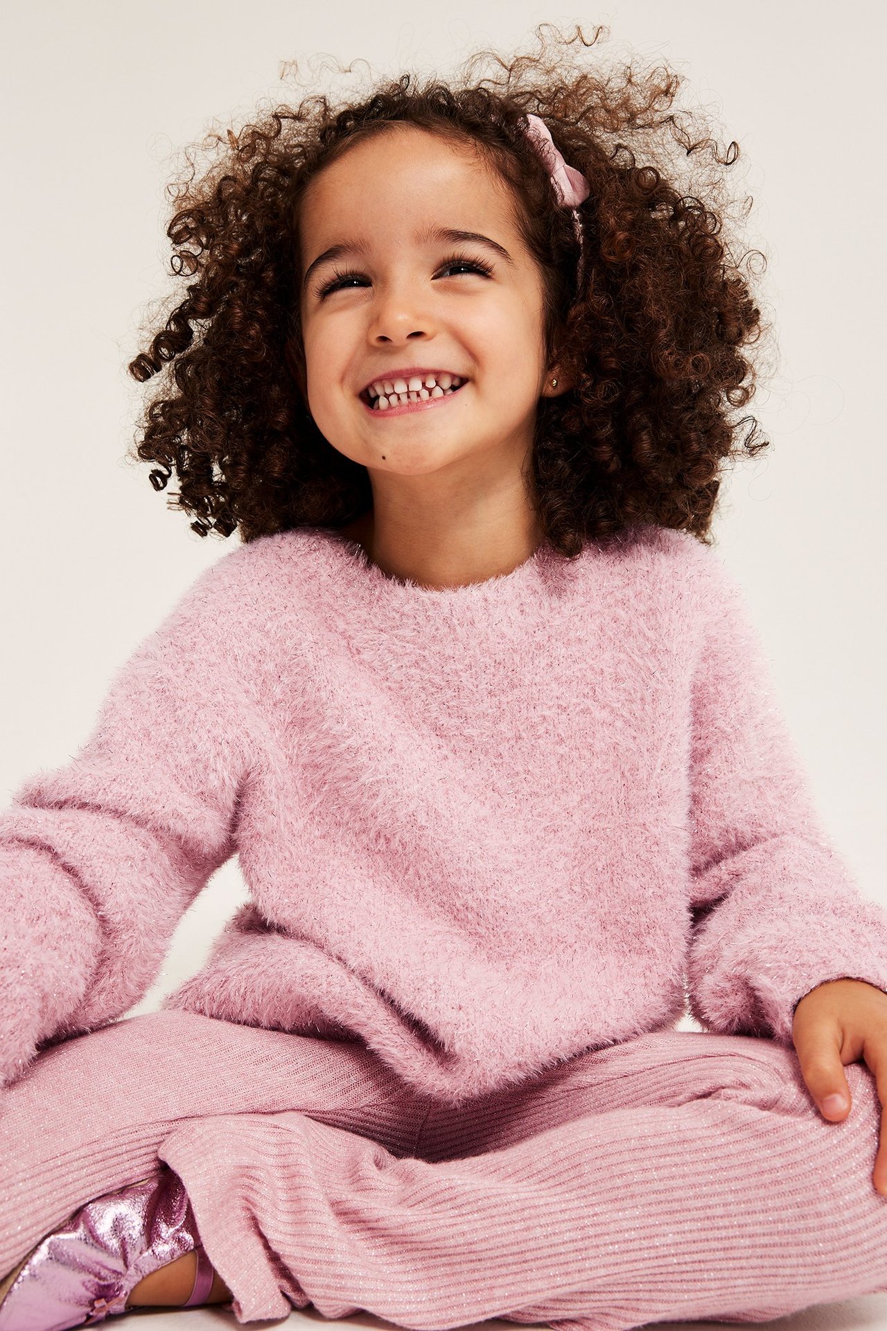 Przód: Uśmiechnięte dziecko w jasnoróżowym puszystym swetrze z brokatem, pasujących spodniach w prążek i błyszczących butach.