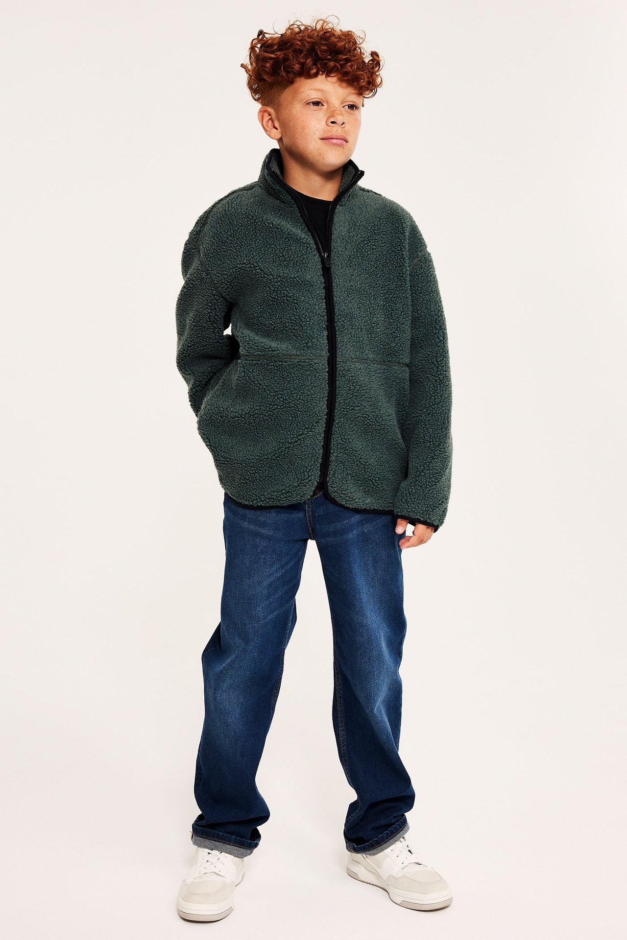 Framifrån: ett barn i en oversized khakigrön pilejacka, mörkblå jeans och vita sneakers.