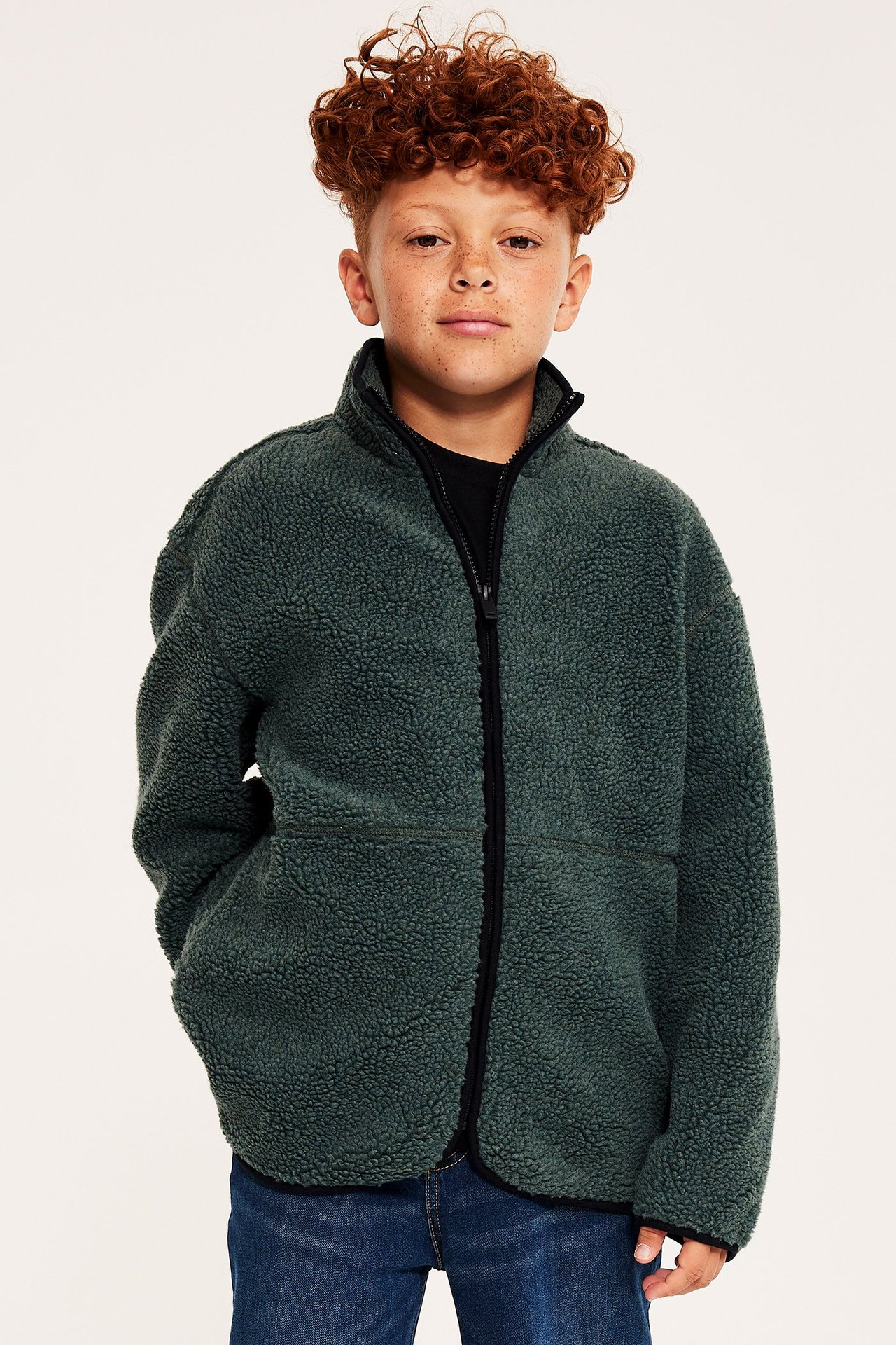 Dziecko w oversize'owej kurtce polarowej khaki z czarnym zamkiem, na czarnym T-shircie i niebieskich dżinsach.