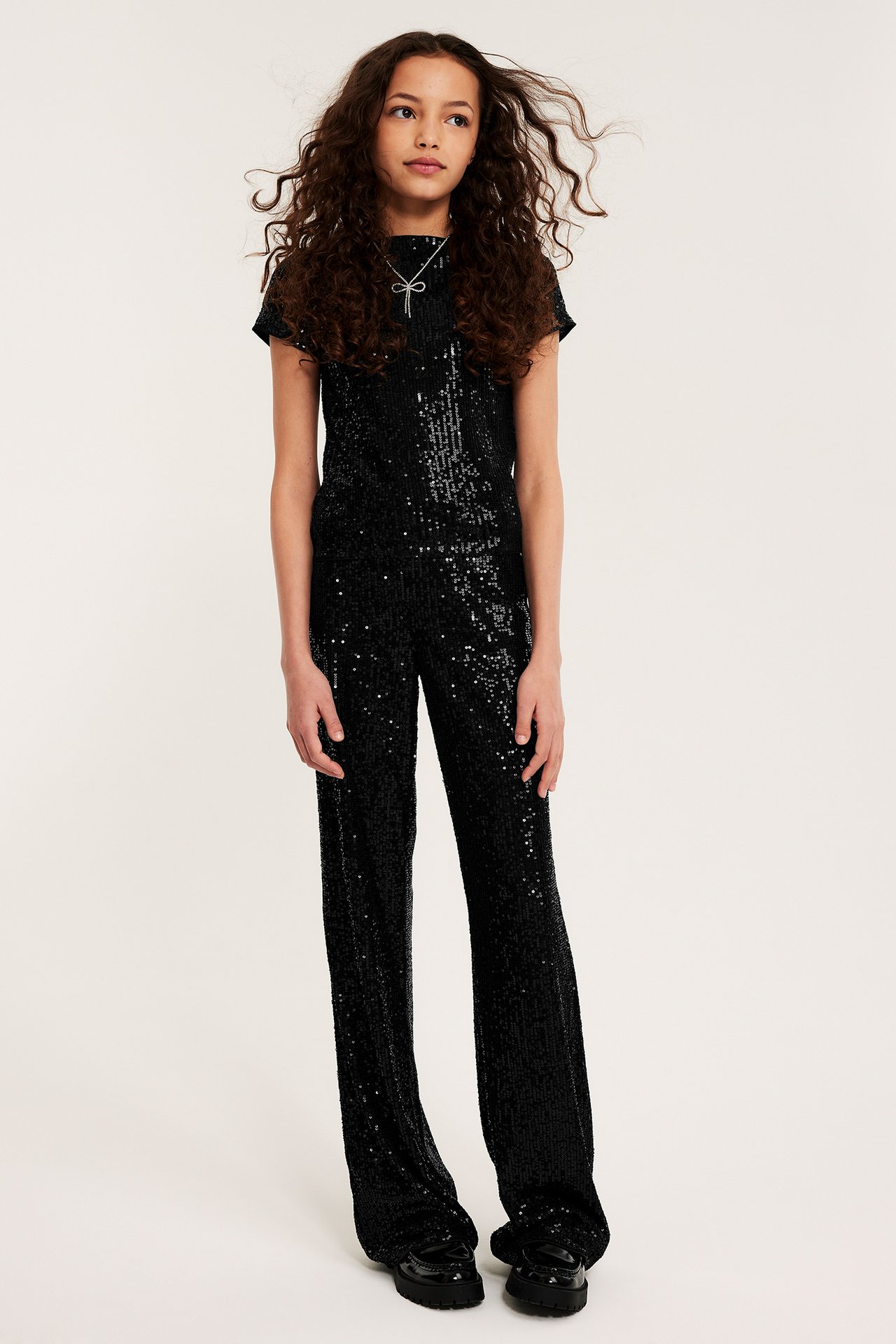 Framifrån: kid i svarta paljettbyxor med vida ben, matchande topp, silverrosett-halsband och chunky loafers.