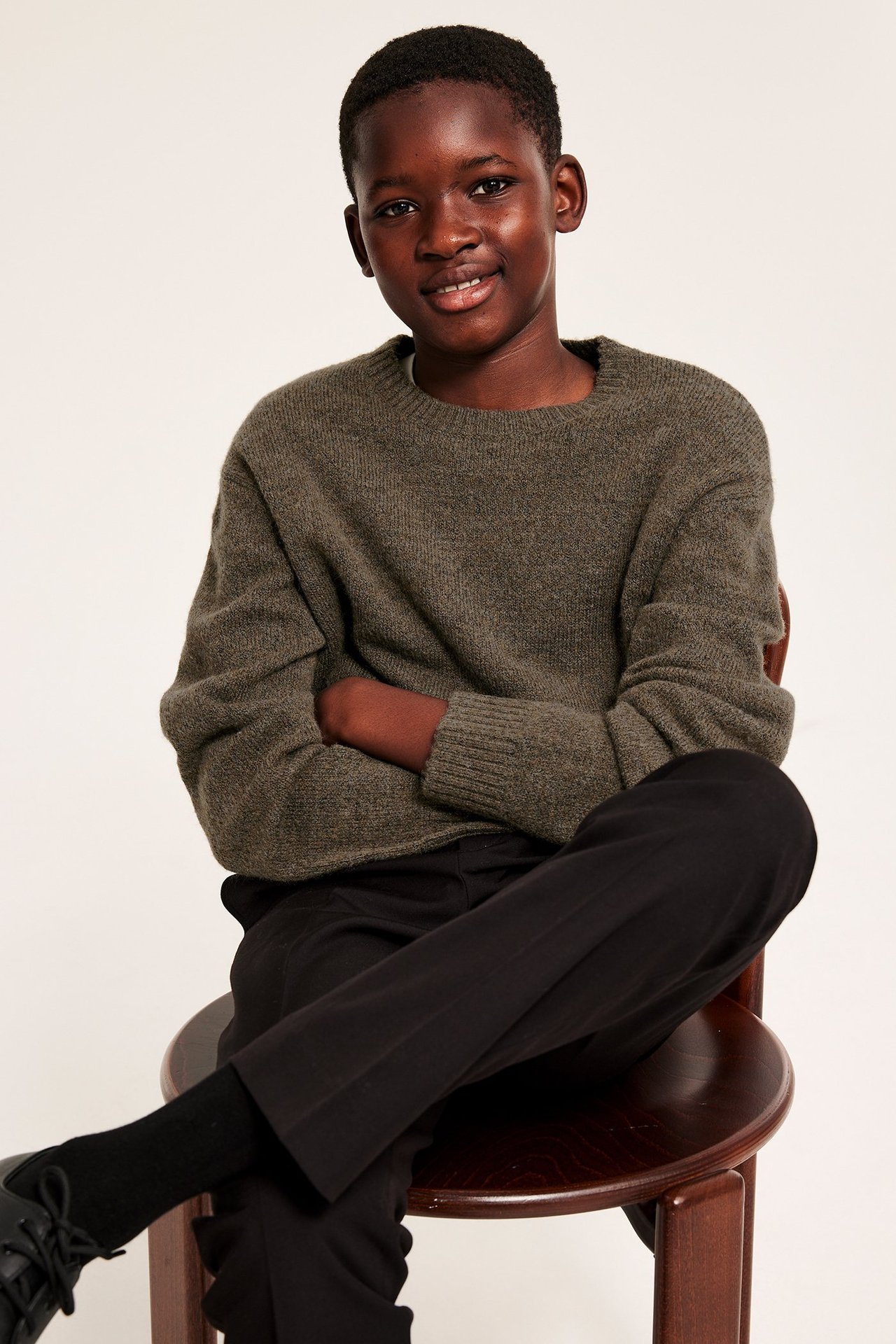 Edestäpäin kuva lapsesta, yllään ruskea neulepaita, mustat housut ja mustat nauhakengät.