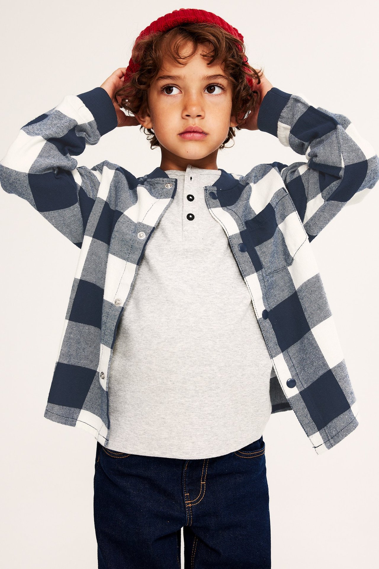 Framifrån: Barn i mörkblå och vitrutig flanellskjorta, grå t-shirt, mörkblå jeans, röd mössa.