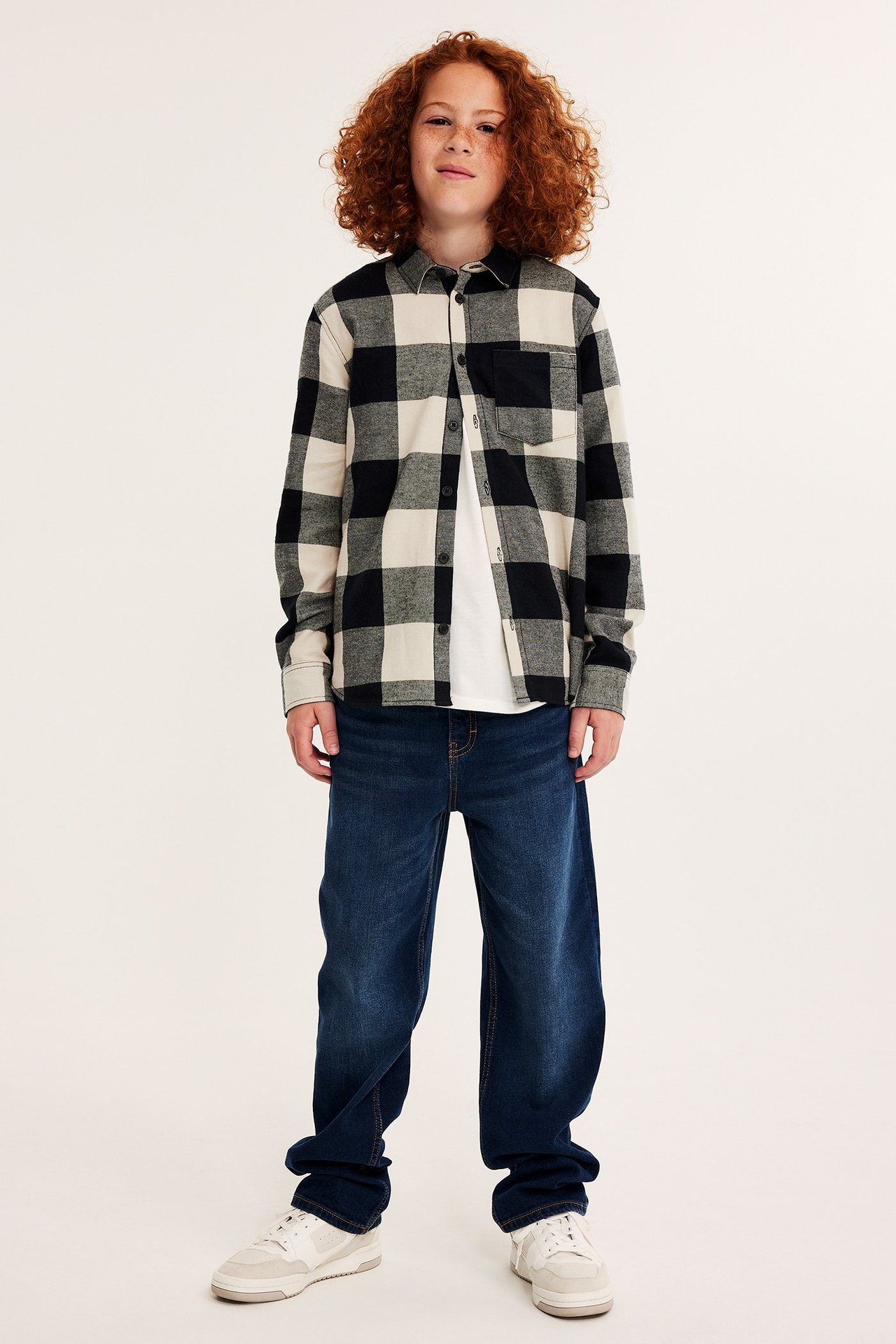 Framifrån: Barn i svart- och vitrutig flanellskjorta, vit t-shirt, mörkblå jeans och vita sneakers.