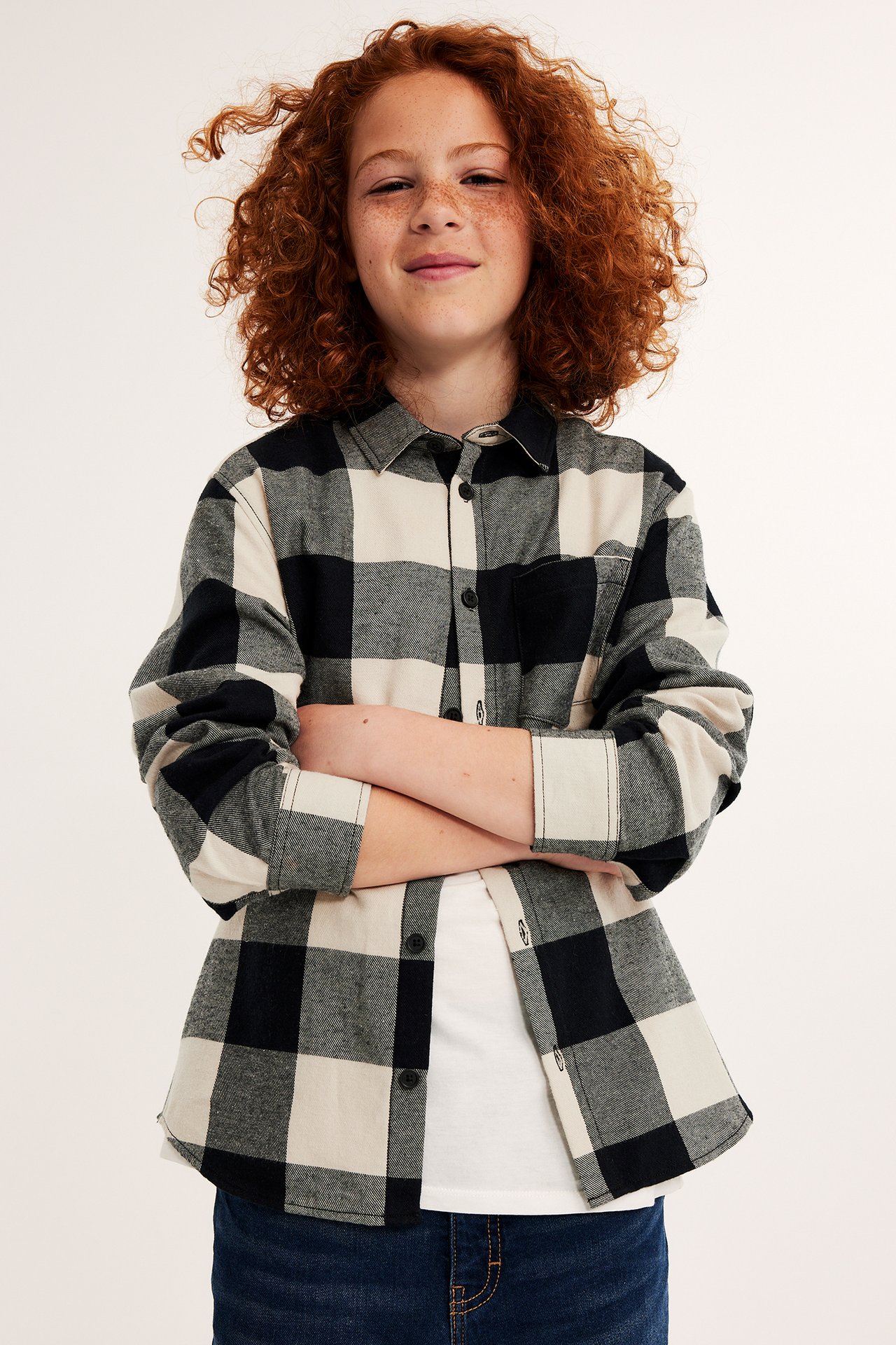 Frontvy: Barn i öppen svart och benvit rutig flanellskjorta över vit t-shirt och mörkblå jeans.