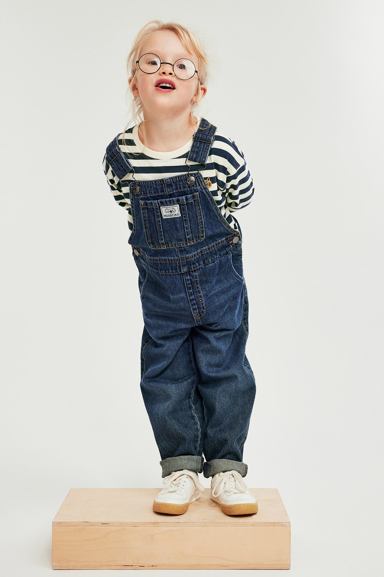 Kid i randig långärmad tröja, mörkblå denimoverall, glasögon och vita sneakers.