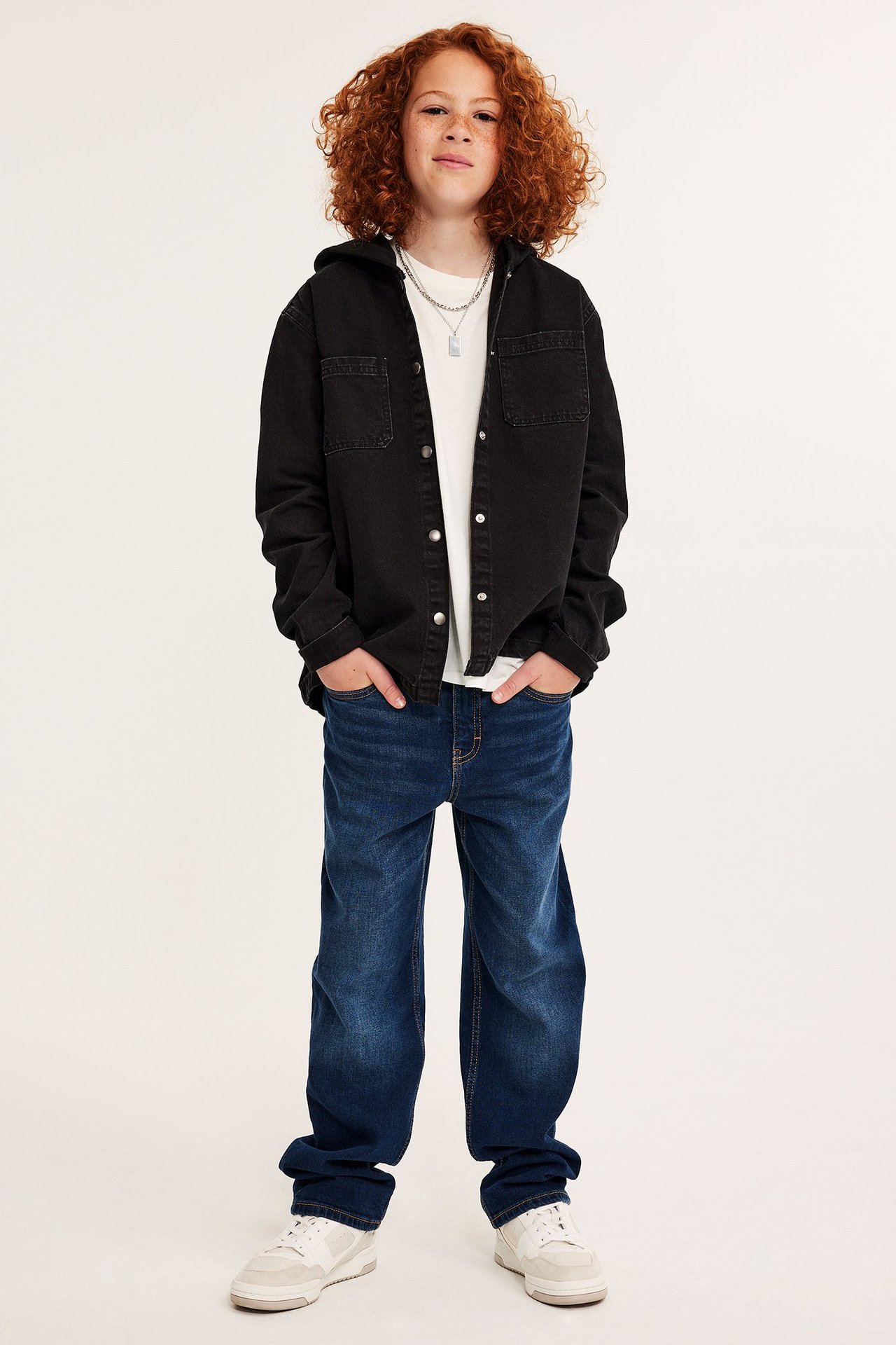 Framifrån: Ett barn i svart jeansskjorta med huva, vit t-shirt, blå jeans och vita sneakers.