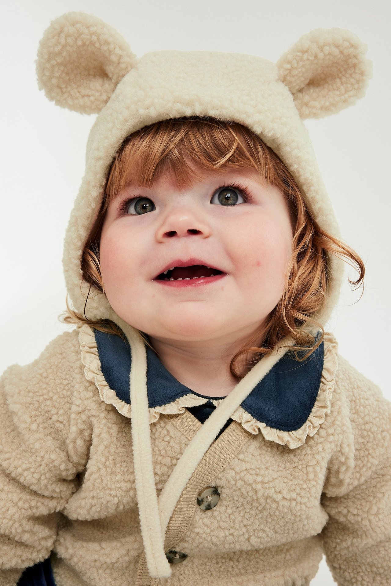 Beige pileliivi nappille, pieni vauva hattussa ja fleece takissa.