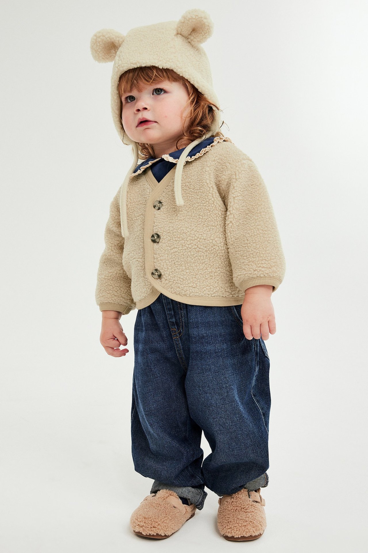 Beige pilekofta med knappar på liten flicka, bär jeans, beige tofflor och mössa med öron.