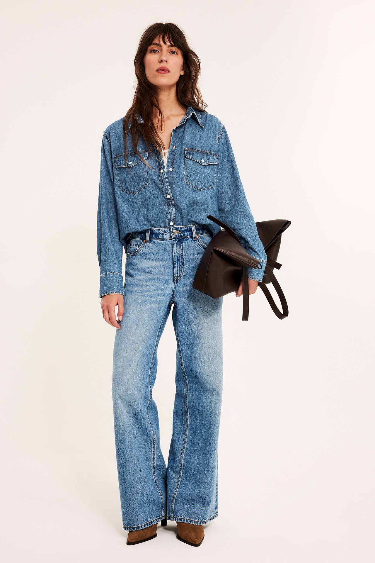 Nainen yllään sininen farkkupaita, vaaleansiniset farkut ja ruskeat saappaat, pitäen ruskeaa käsilaukkua.