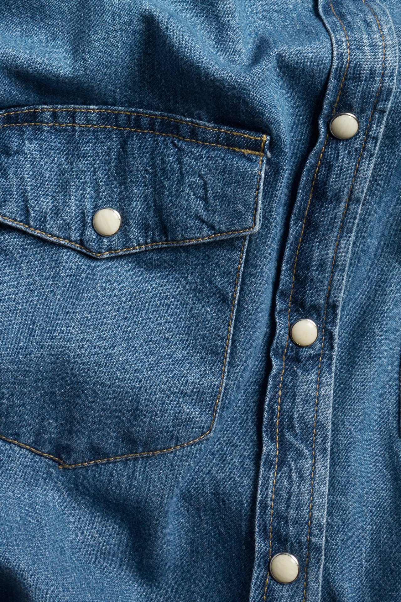 Zbliżenie niebieskiej koszuli jeansowej z kieszenią na piersi, klapką i jasnymi guzikami na plisie.