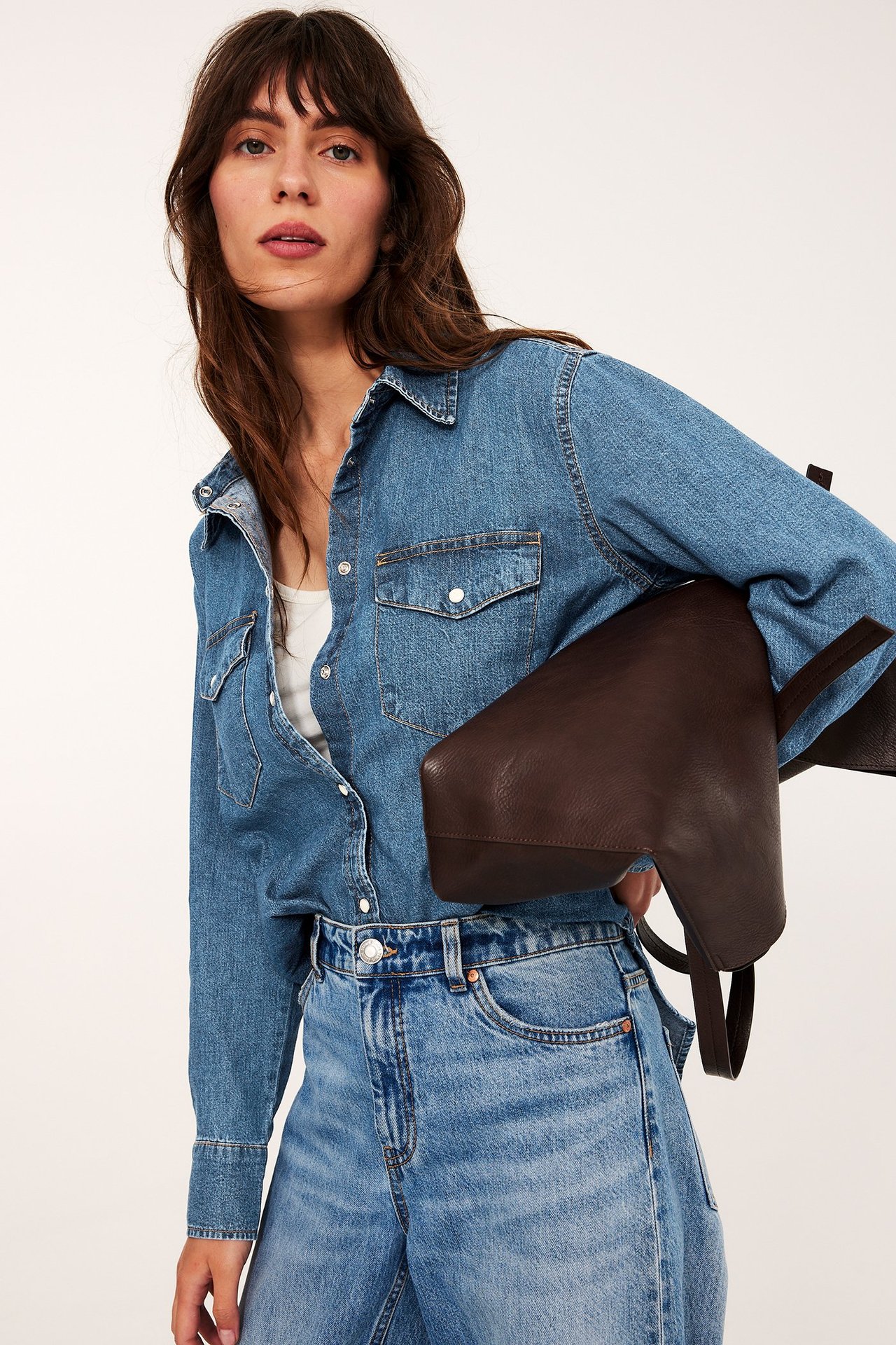 Widok z przodu: Kobieta w otwartej jasnoniebieskiej koszuli jeansowej, białym topie, jasnoniebieskich jeansach, z ciemnobrązową torbą.