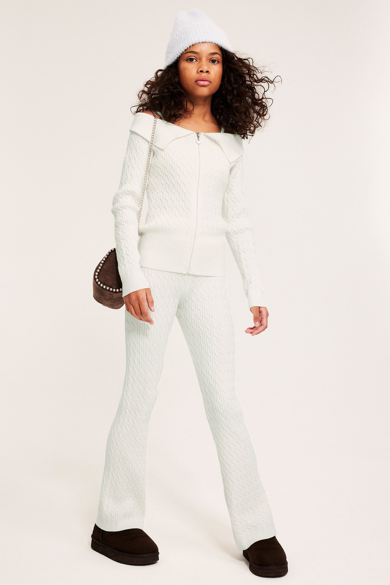 Widok z przodu dziecka w écru dzianinowych spodniach, pasującym kardiganie, białej czapce, brązowej torbie i ciemnych butach.