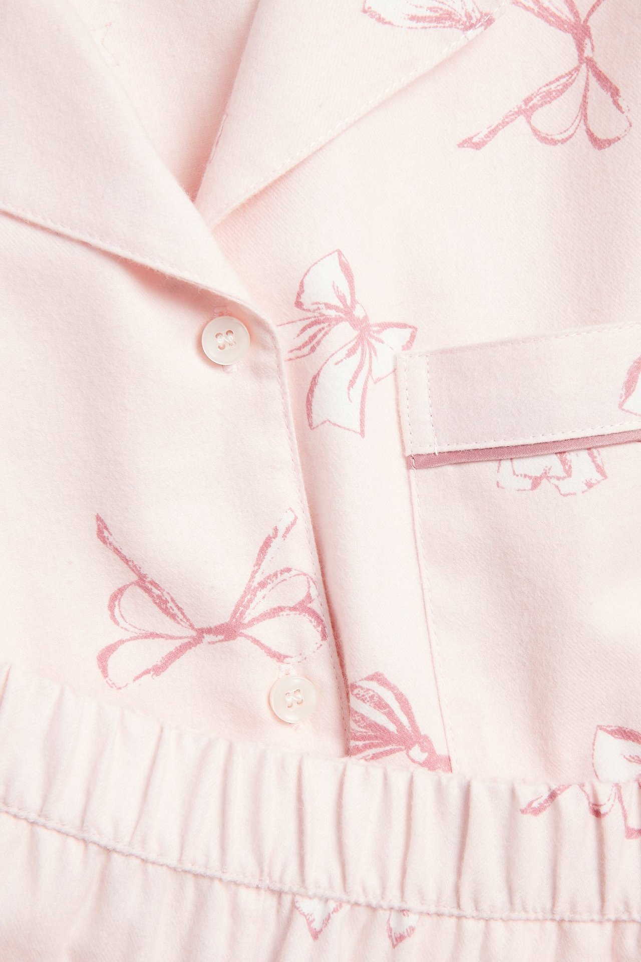 Nærbilde: rosa dame flanellpyjamasoverdel med rosa sløyfemønster, krage, knapper og brystlomme.