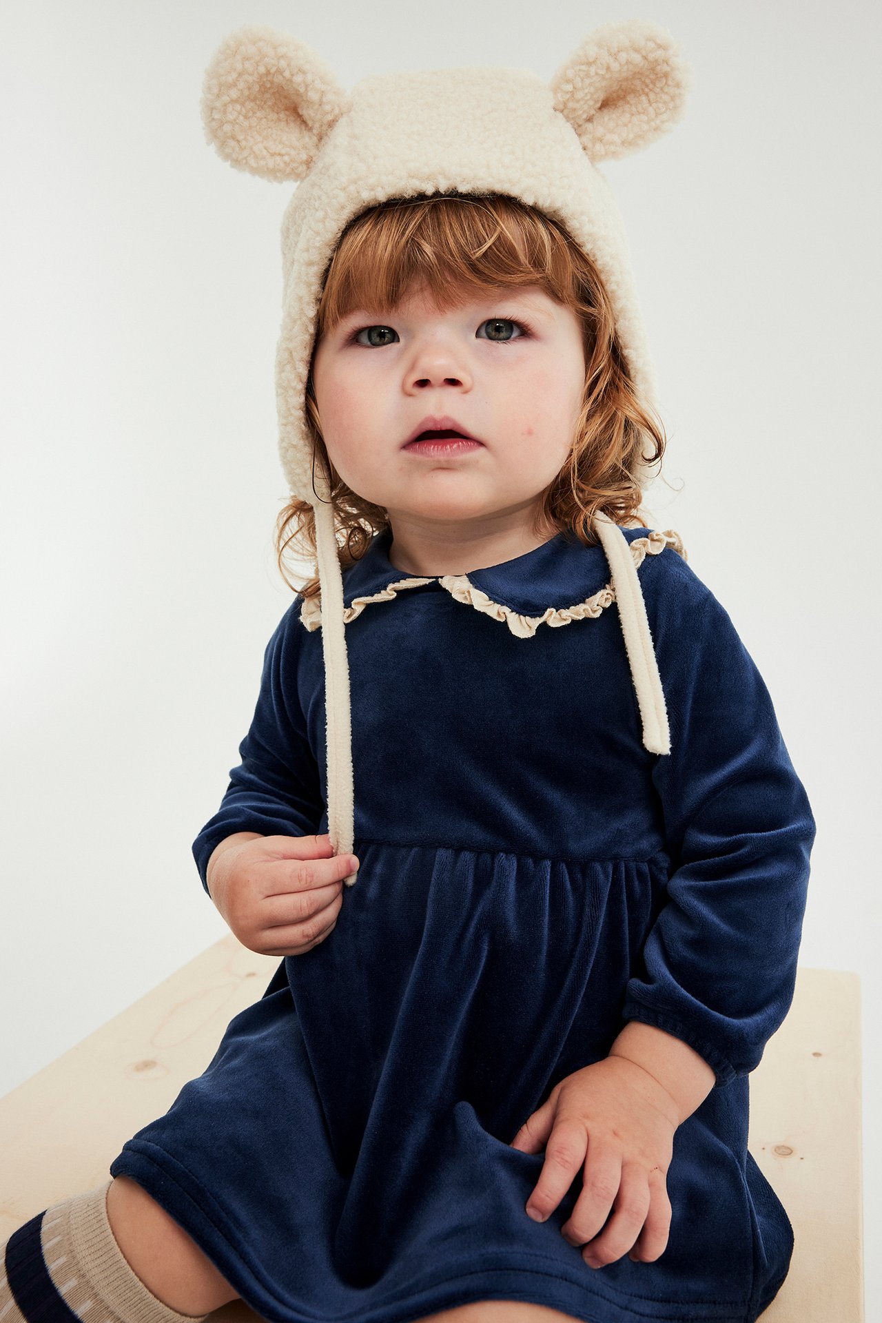 Framifrån, en baby i en beige pilémössa med öron, en mörkblå sammetsklänning och randiga strumpor.
